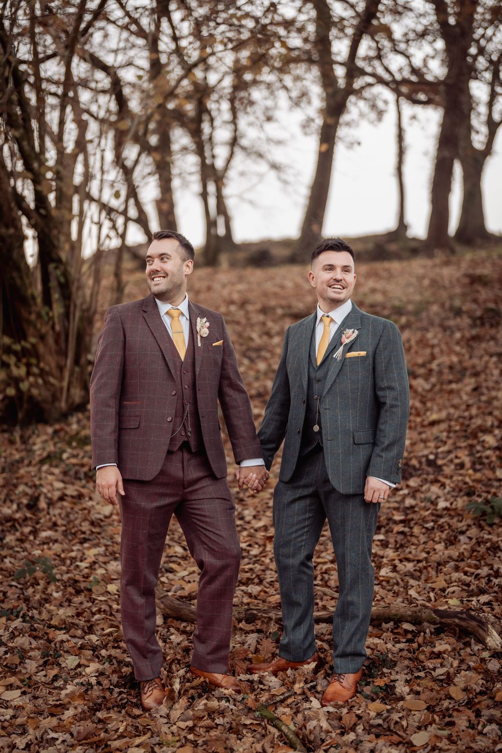 Two people in complementary suits hold hands while standing among autumn leaves and bare trees.