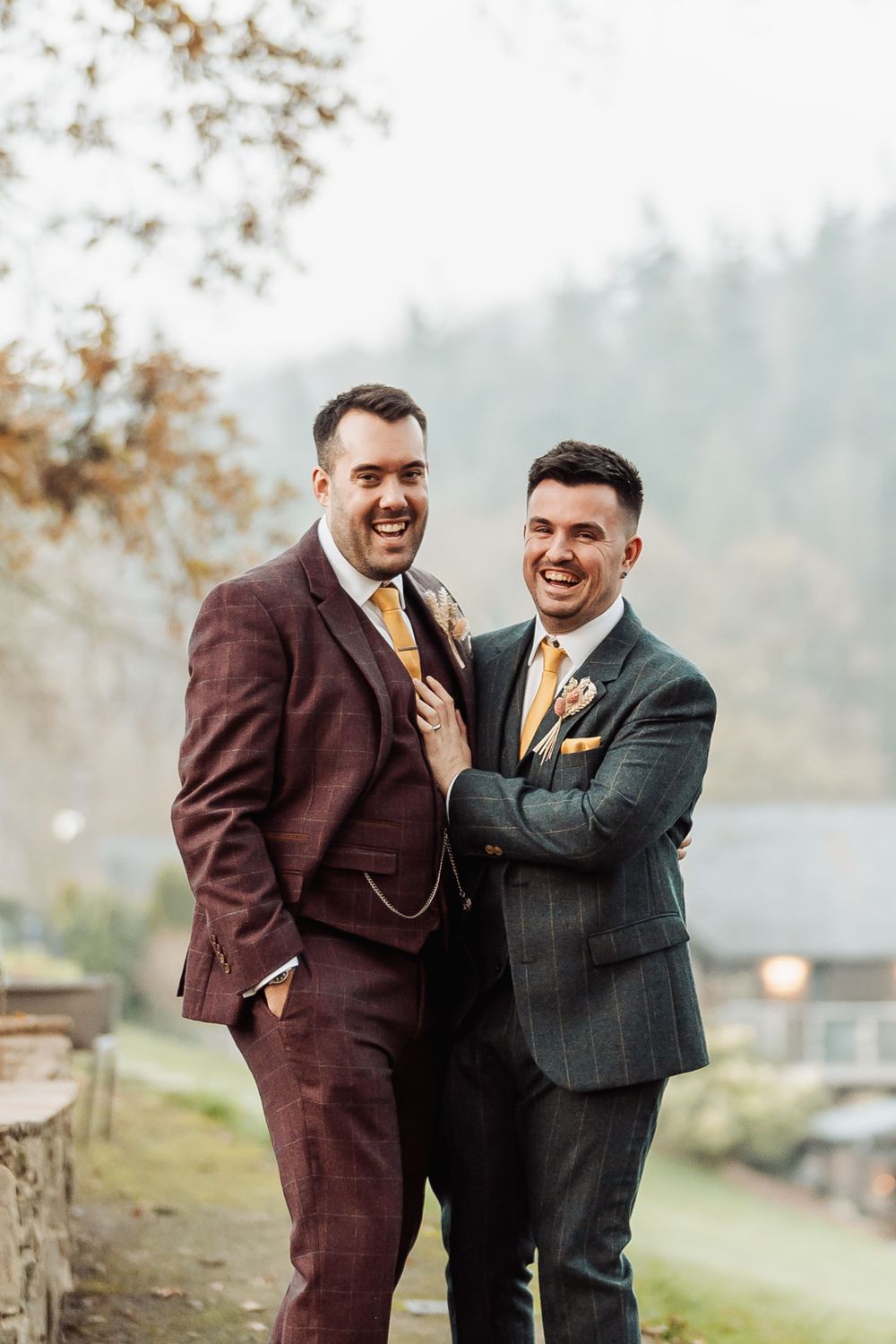 Two people in formal attire share a joyful moment on an autumn path overlooking water.