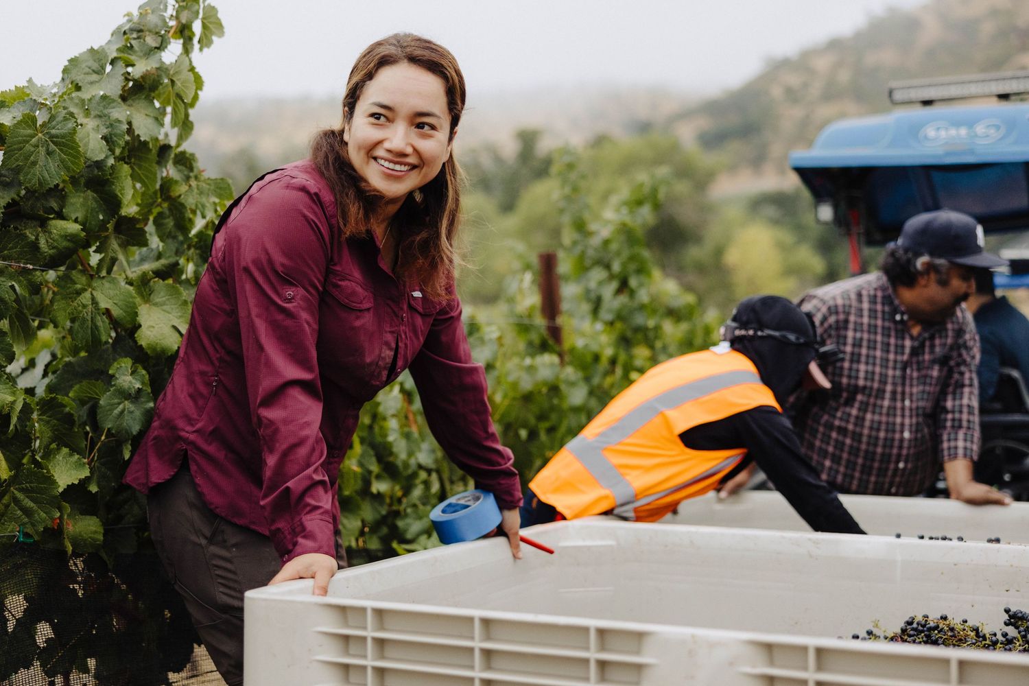 A woman smiles to someone off camera while standing on the back of a tractor in a vineyard in Paso Robles, California
