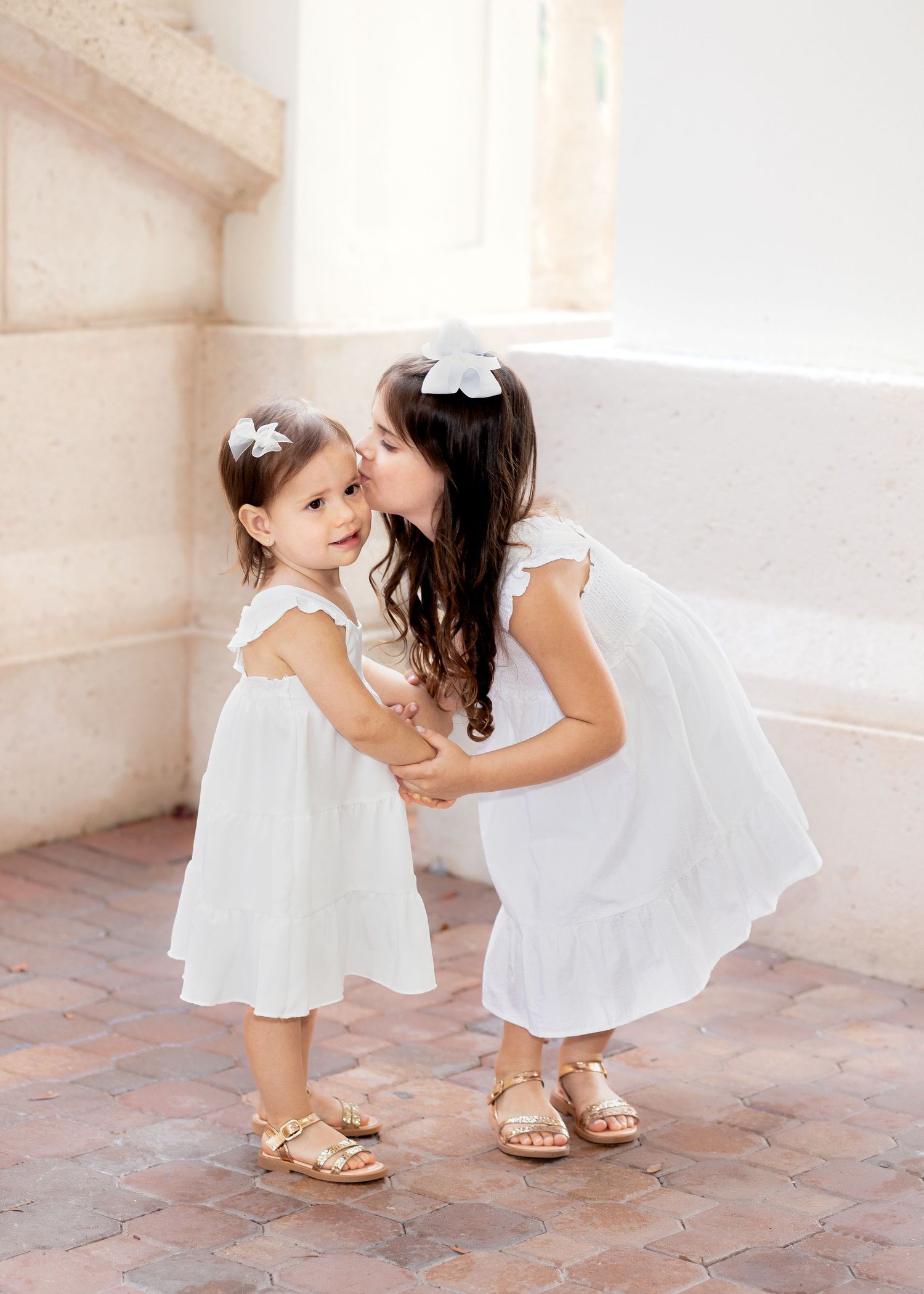 Two children in white dresses share a sweet moment together wearing matching hair bows and sandals.