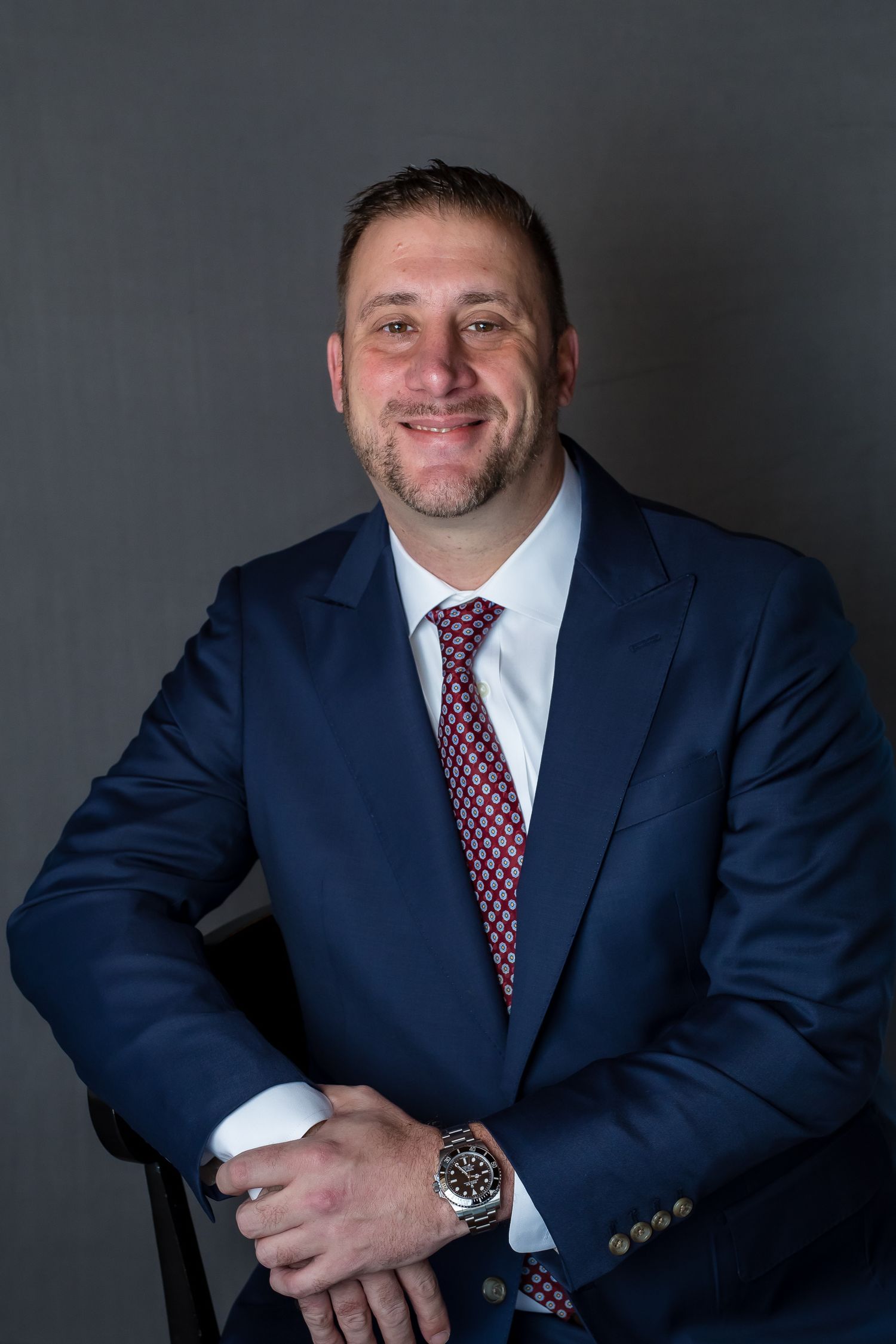 Professional headshot with navy suit and red patterned tie against dark background.