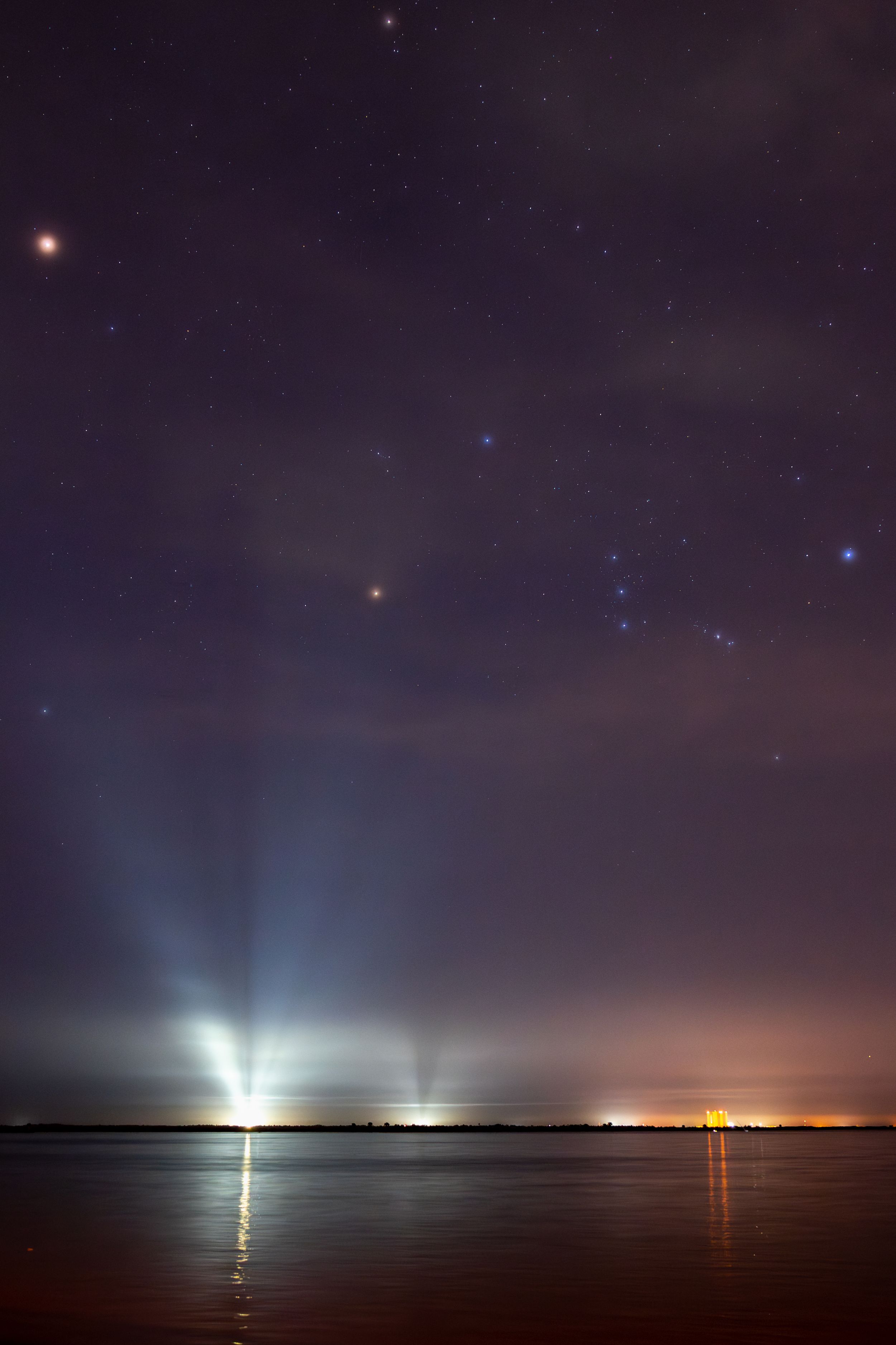 Orion constellation, and Planet Mars just above the Artemis SLS Night sky over water with stars and light beams reflecting on the surface.
