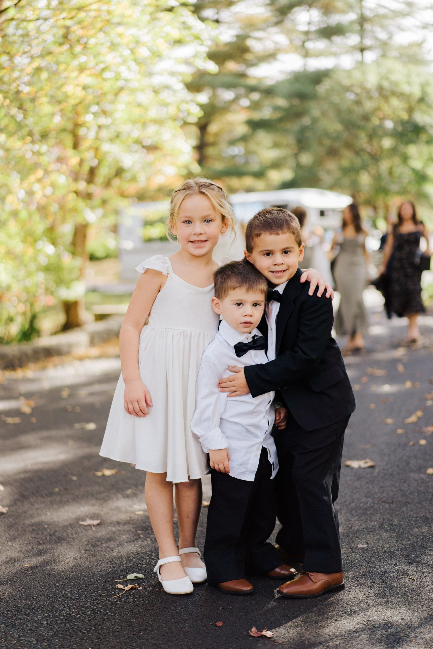 Three young children in formal attire pose together on a paved path surrounded by autumn foliage.