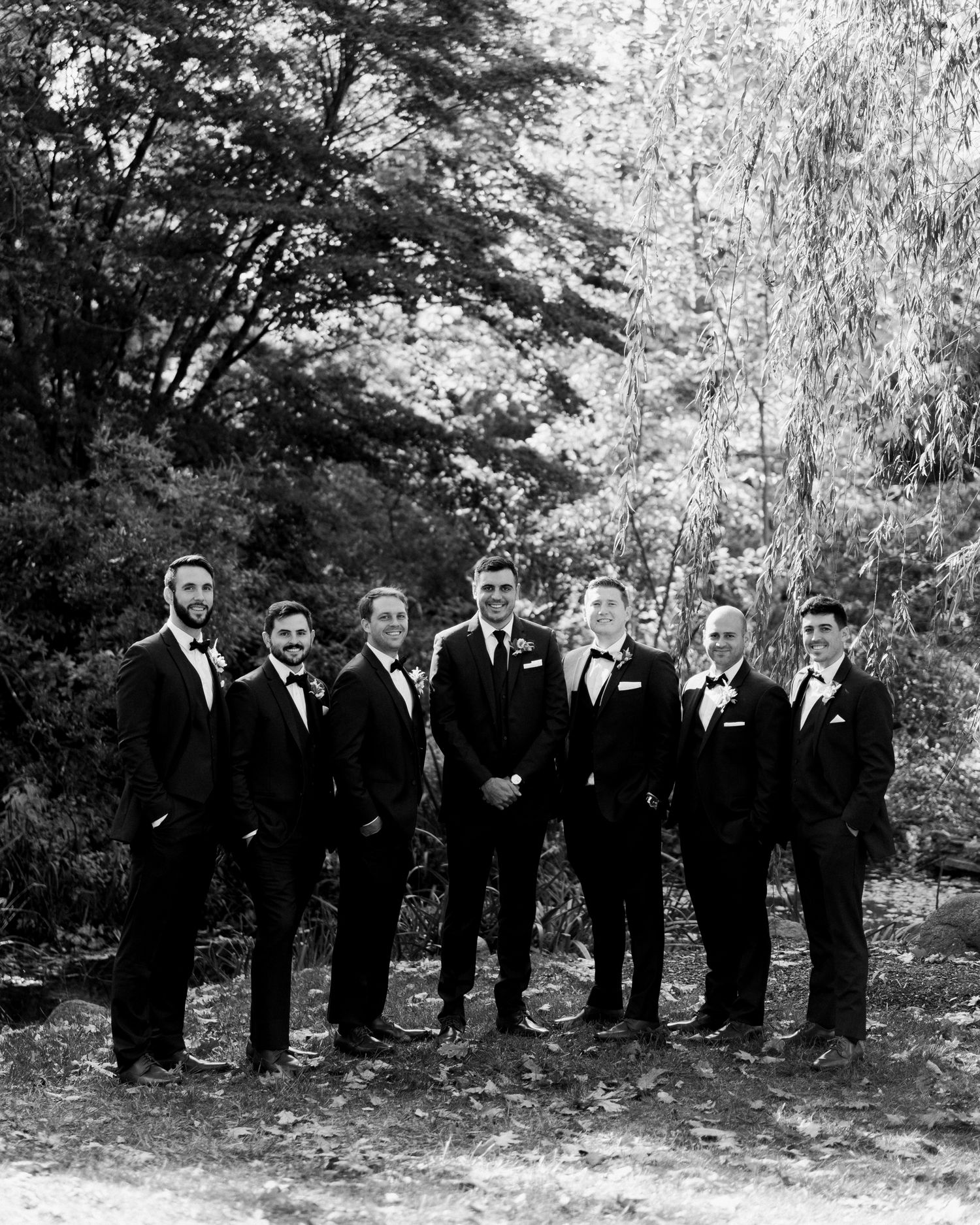 Groomsmen in black suits pose together against natural foliage background in black and white photography.