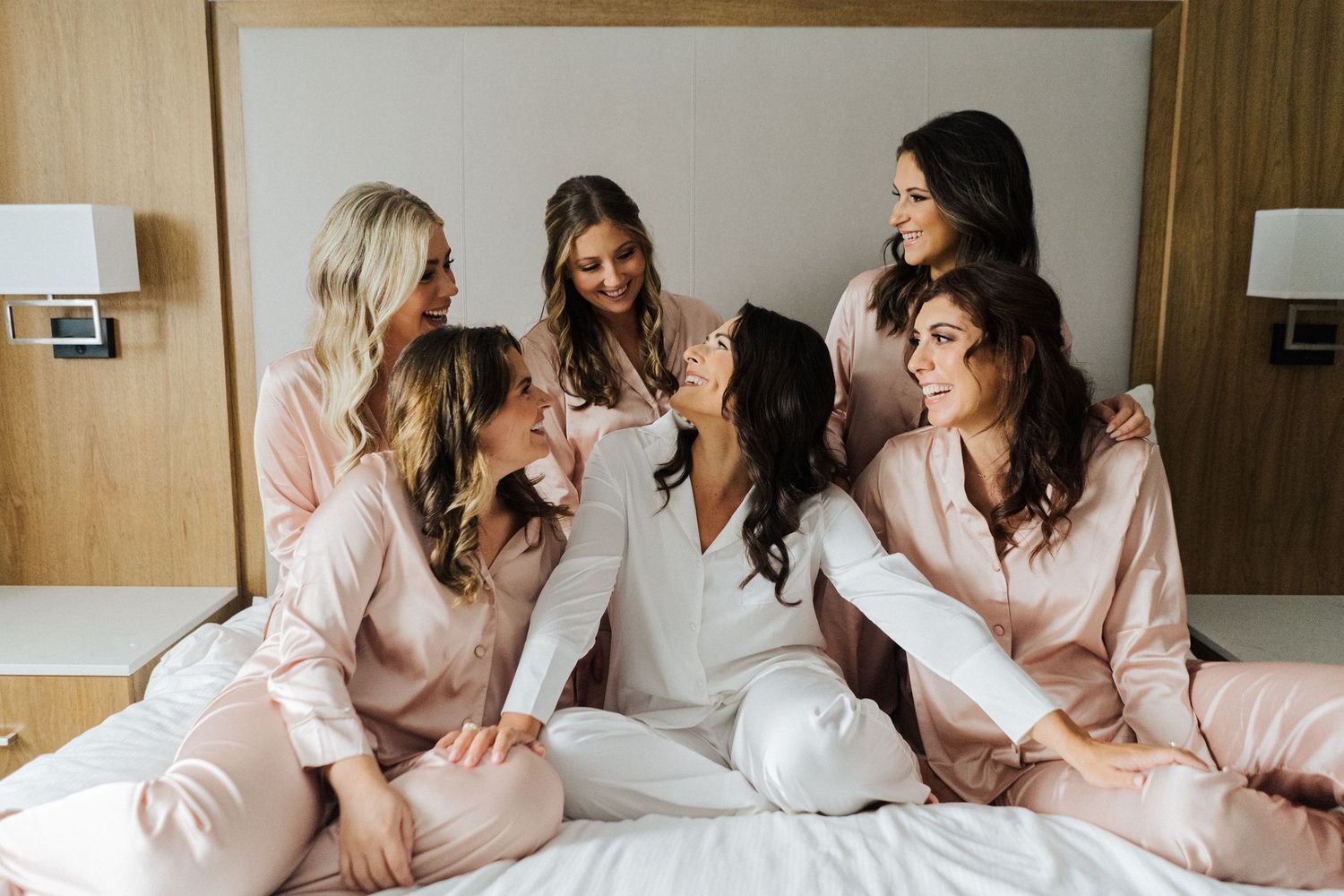 Bridal party in blush pink robes gather on hotel bed for pre-wedding photos.