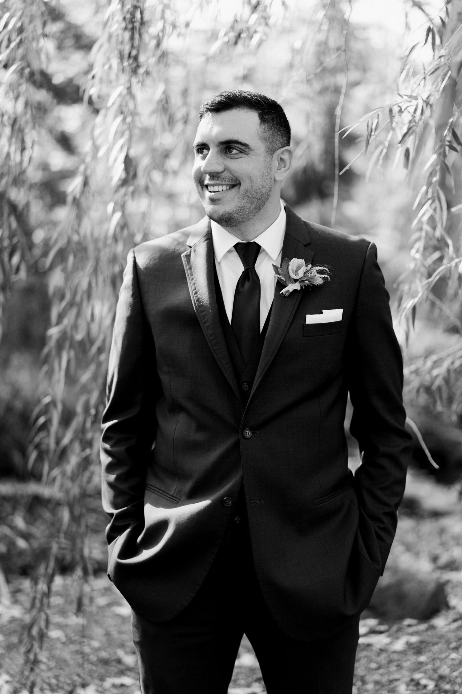 Black and white portrait of groom in classic suit under willow branches.