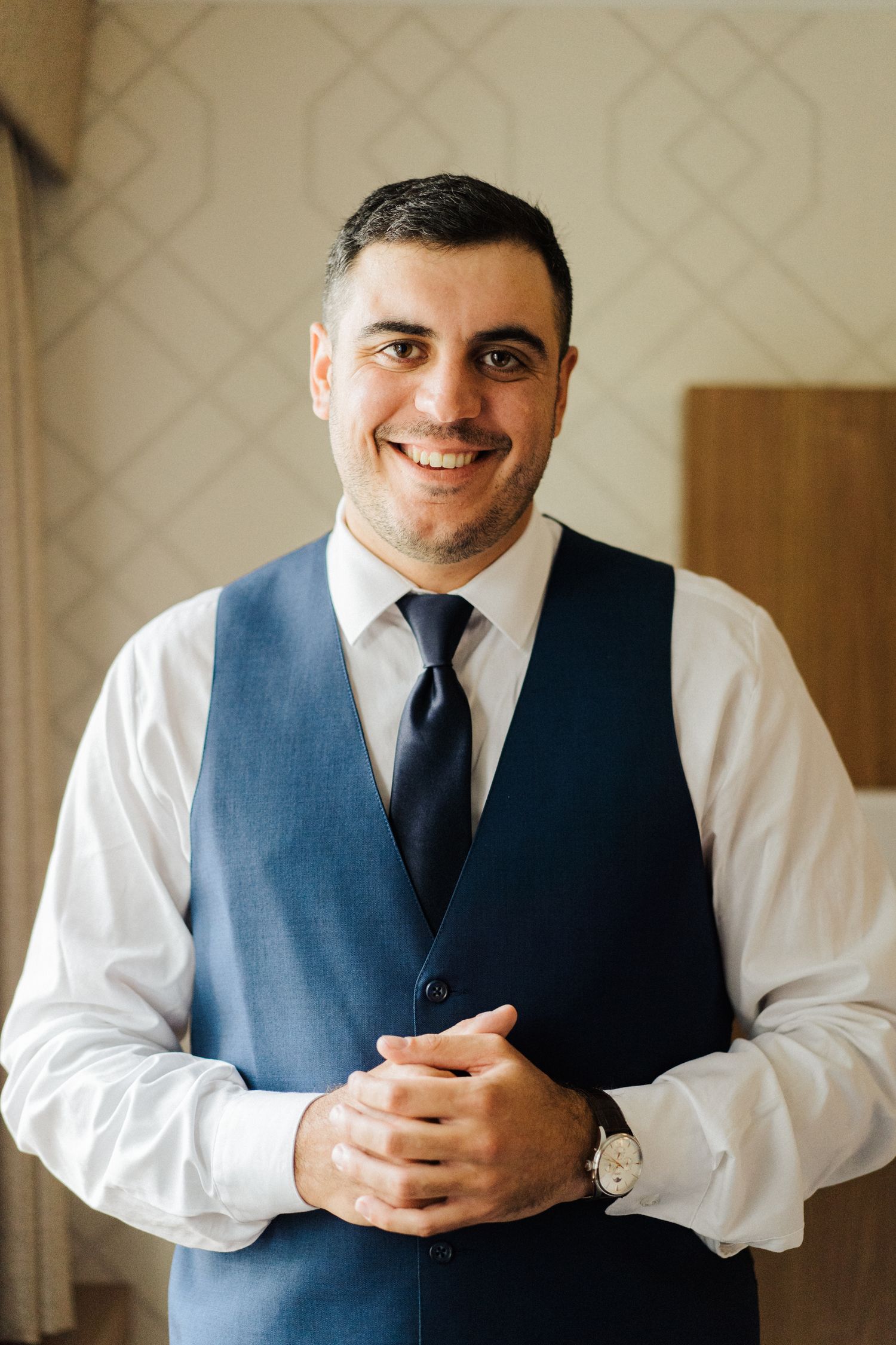 Portrait of person in blue vest and tie smiling at camera against patterned wall.