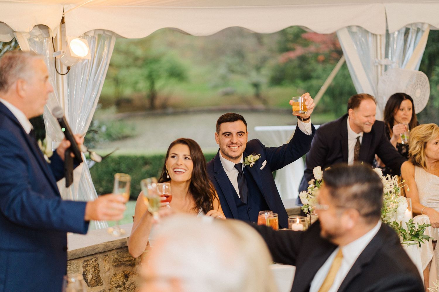 Wedding guests raise their glasses during a celebratory toast at an outdoor tented reception.