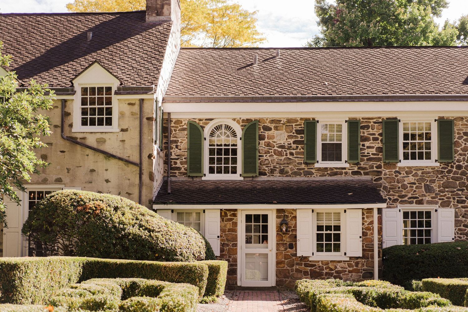 Stone colonial house with white trim windows and manicured hedges in afternoon light.
