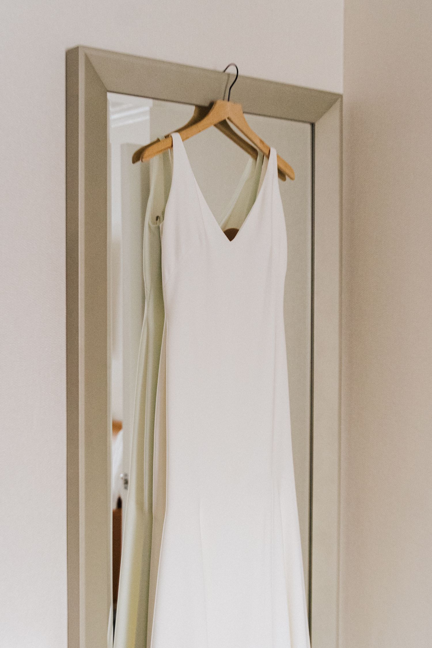 Minimalist white wedding dress hanging on wooden hanger against neutral wall.