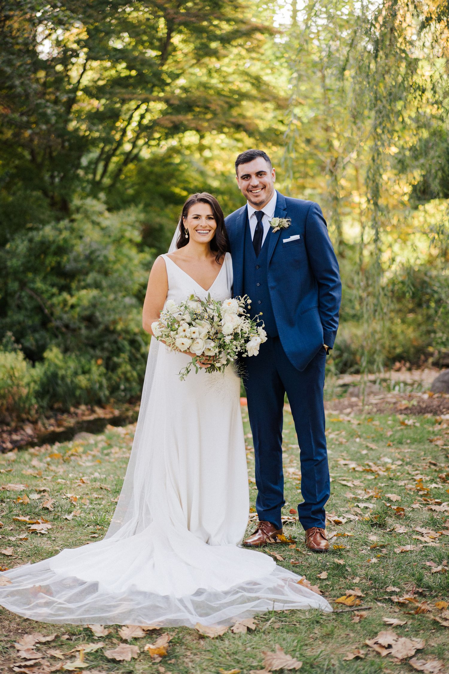 Bride and groom pose outdoors in elegant white dress and navy suit.