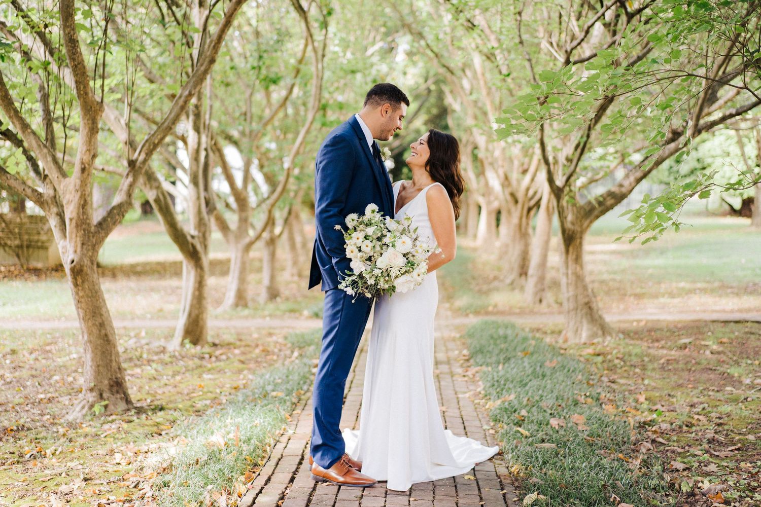 Romantic wedding portrait on brick pathway under sunlit tree canopy.