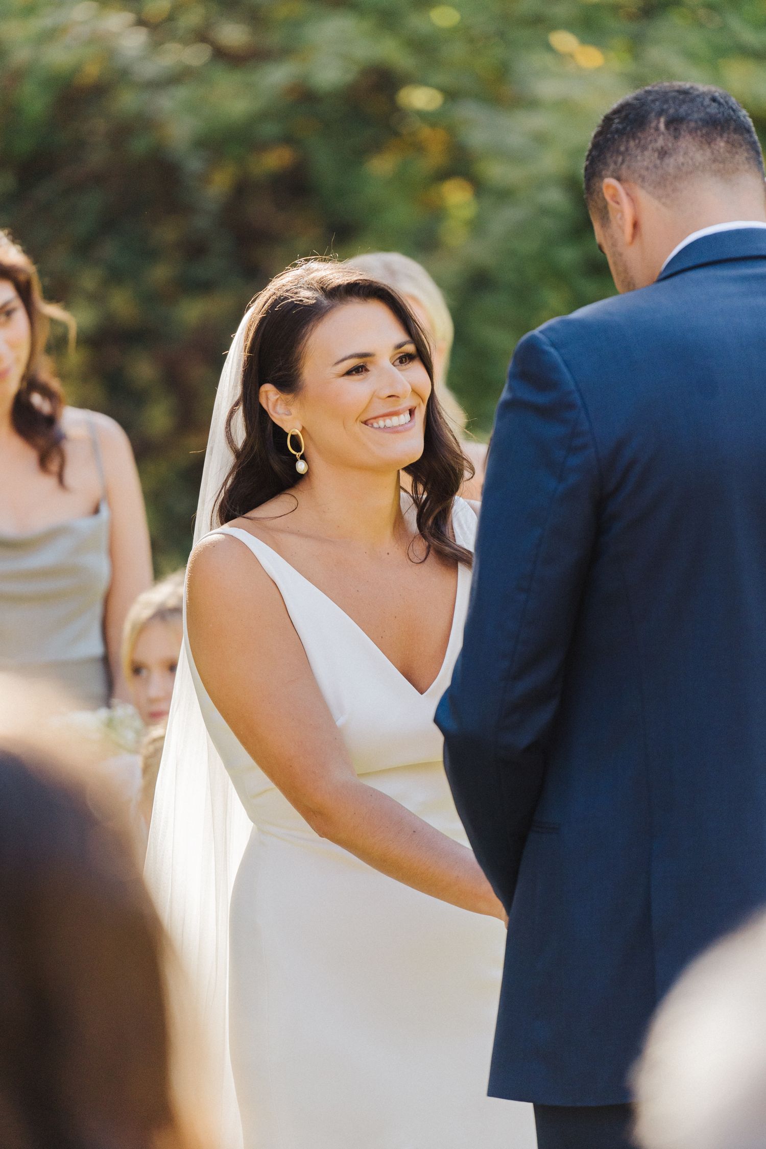 A smiling bride in a sleek white dress exchanges vows during an outdoor ceremony.