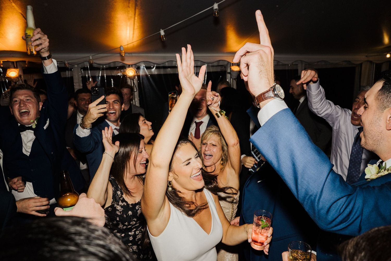 Energetic wedding reception crowd dancing with hands in the air under ambient lighting.