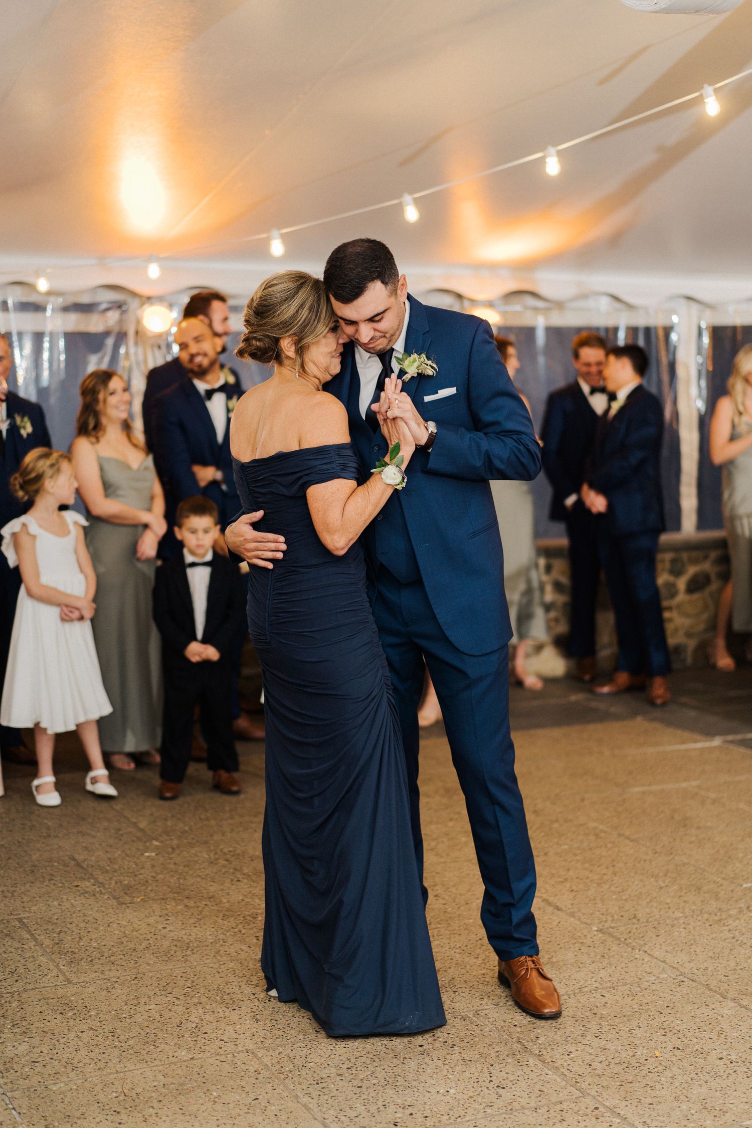 A formal dance between two people in evening attire under string lights at a wedding reception.