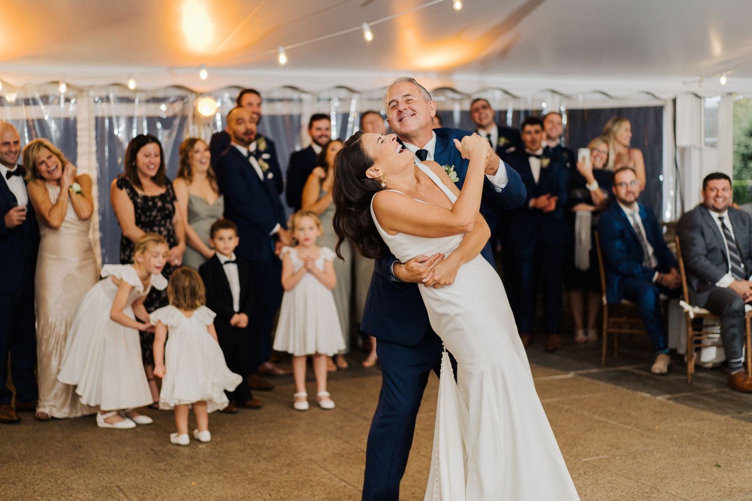 A couple shares their first dance at a wedding reception under a tent while guests watch and smile.