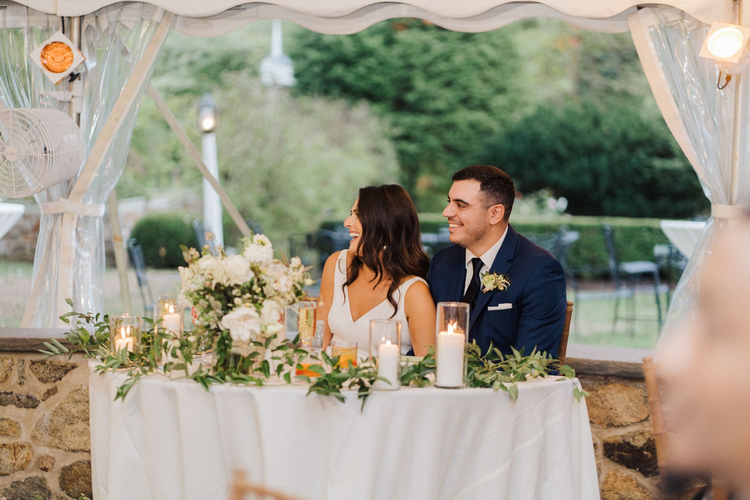 A couple sits at an elegantly decorated sweetheart table under a tent with floral arrangements and candles.