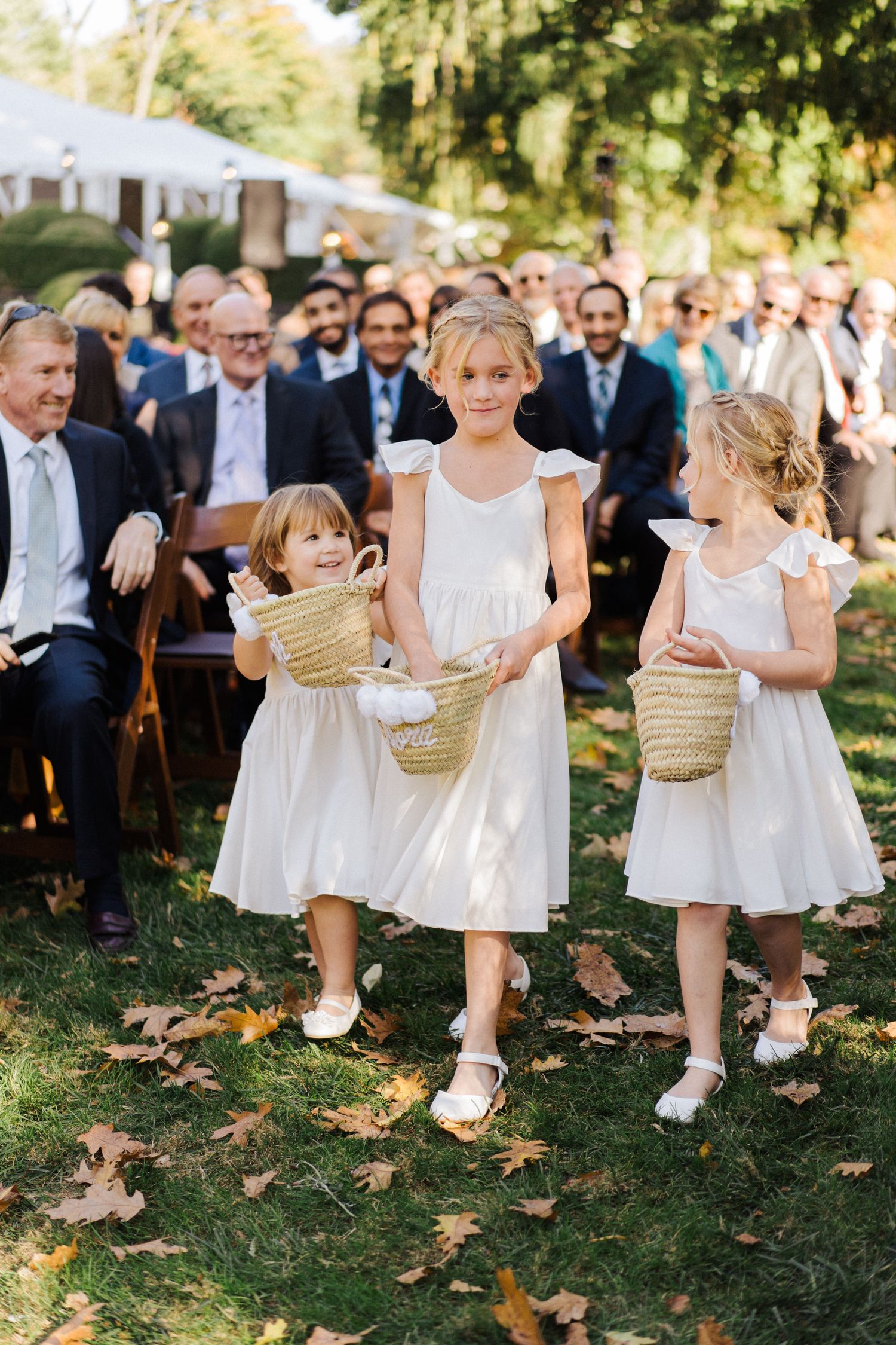 Three young attendants in white dresses carry woven baskets down the aisle at an outdoor wedding ceremony.