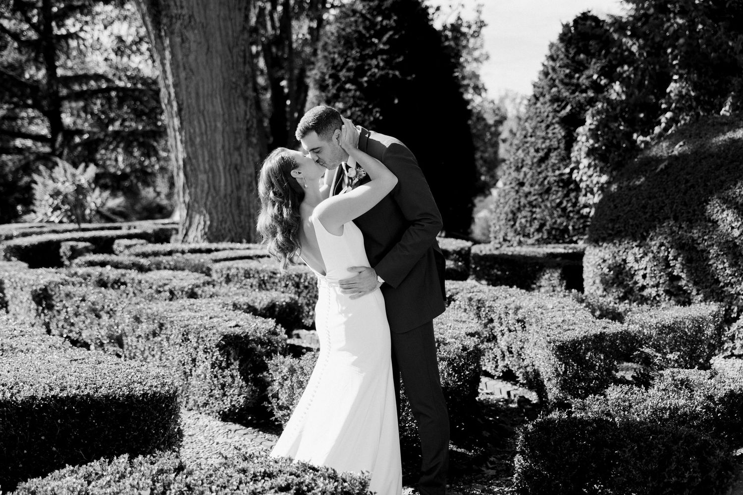 A couple shares a romantic moment in a formal garden setting with manicured hedges in black and white.