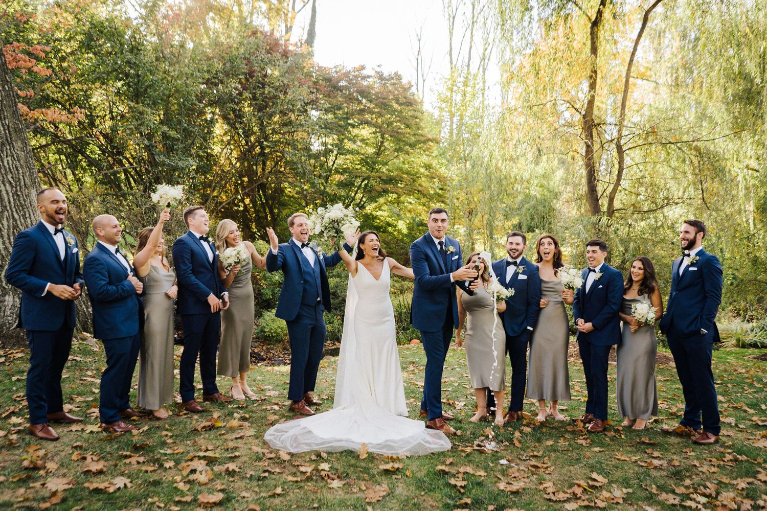 Wedding party celebrates with champagne beneath willow trees during autumn photo session.