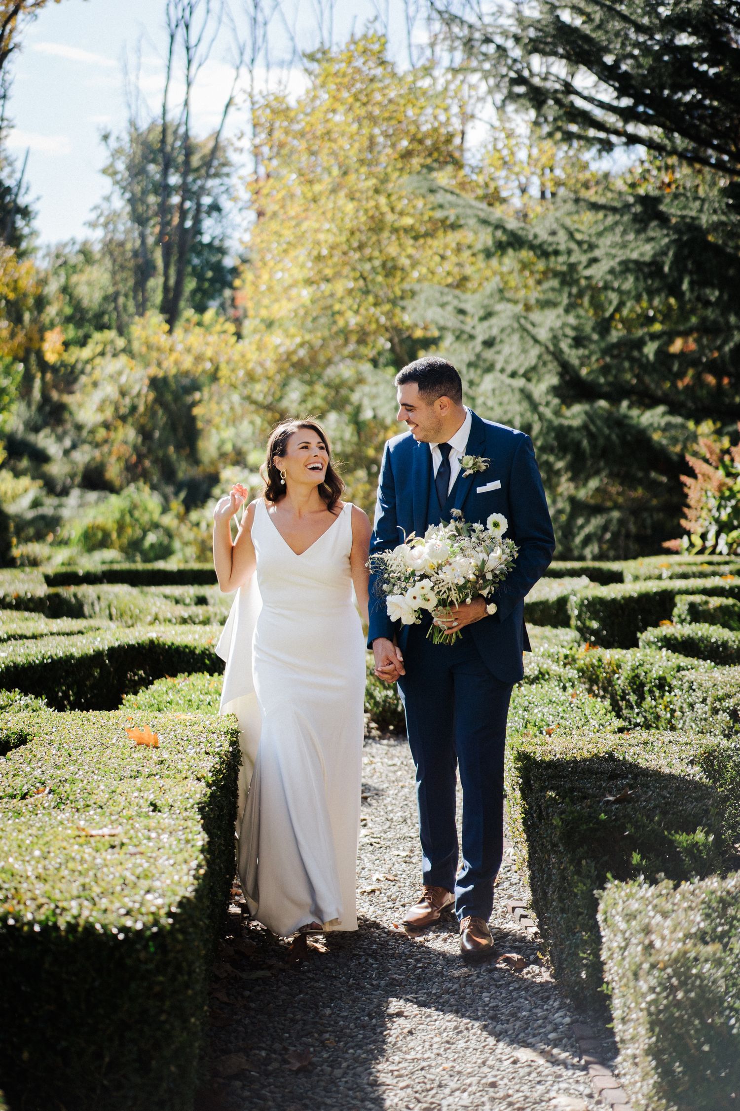Bride and groom walk happily through a formal garden path carrying white floral bouquet.