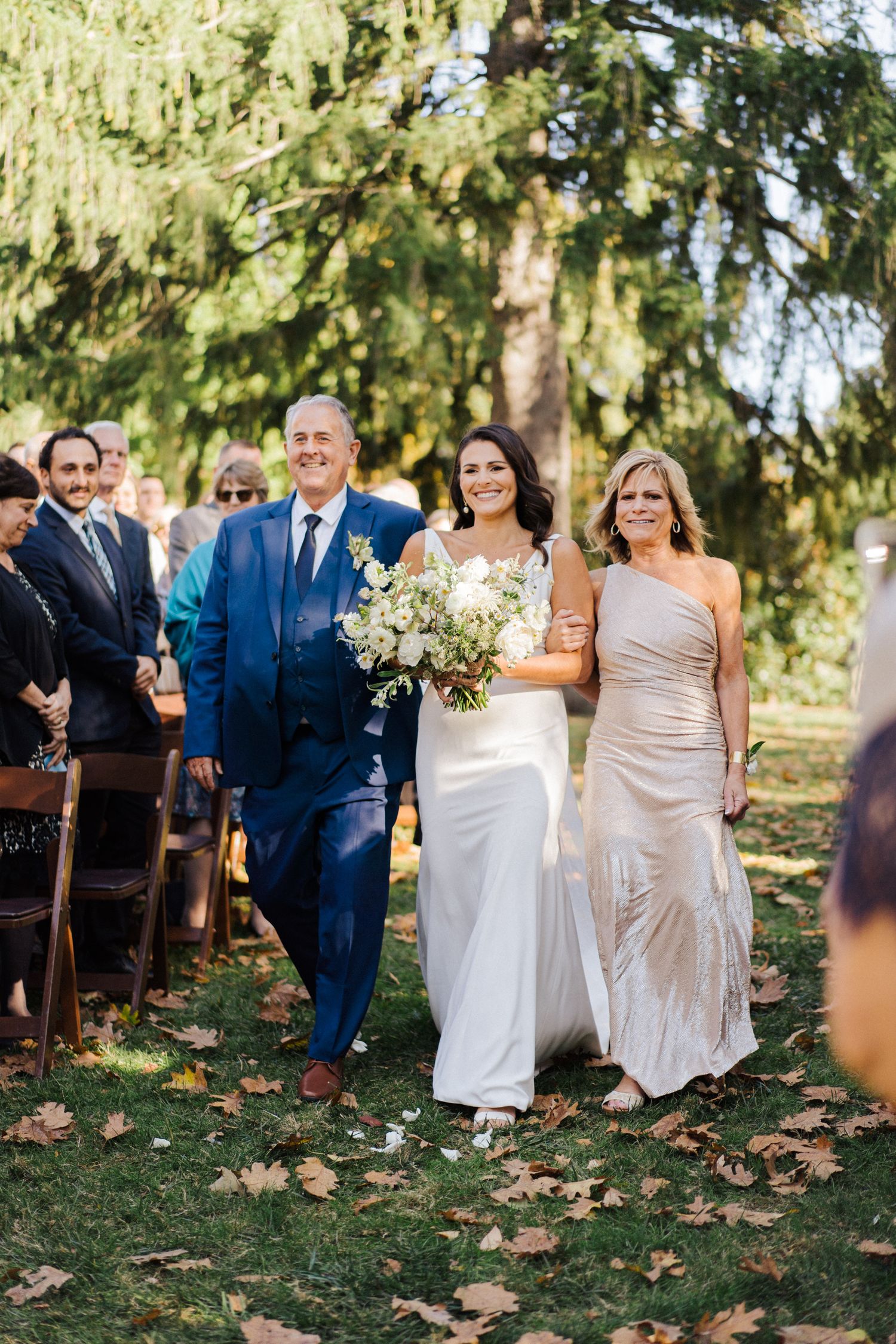Wedding party members walk down an outdoor aisle lined with wooden chairs and autumn leaves.