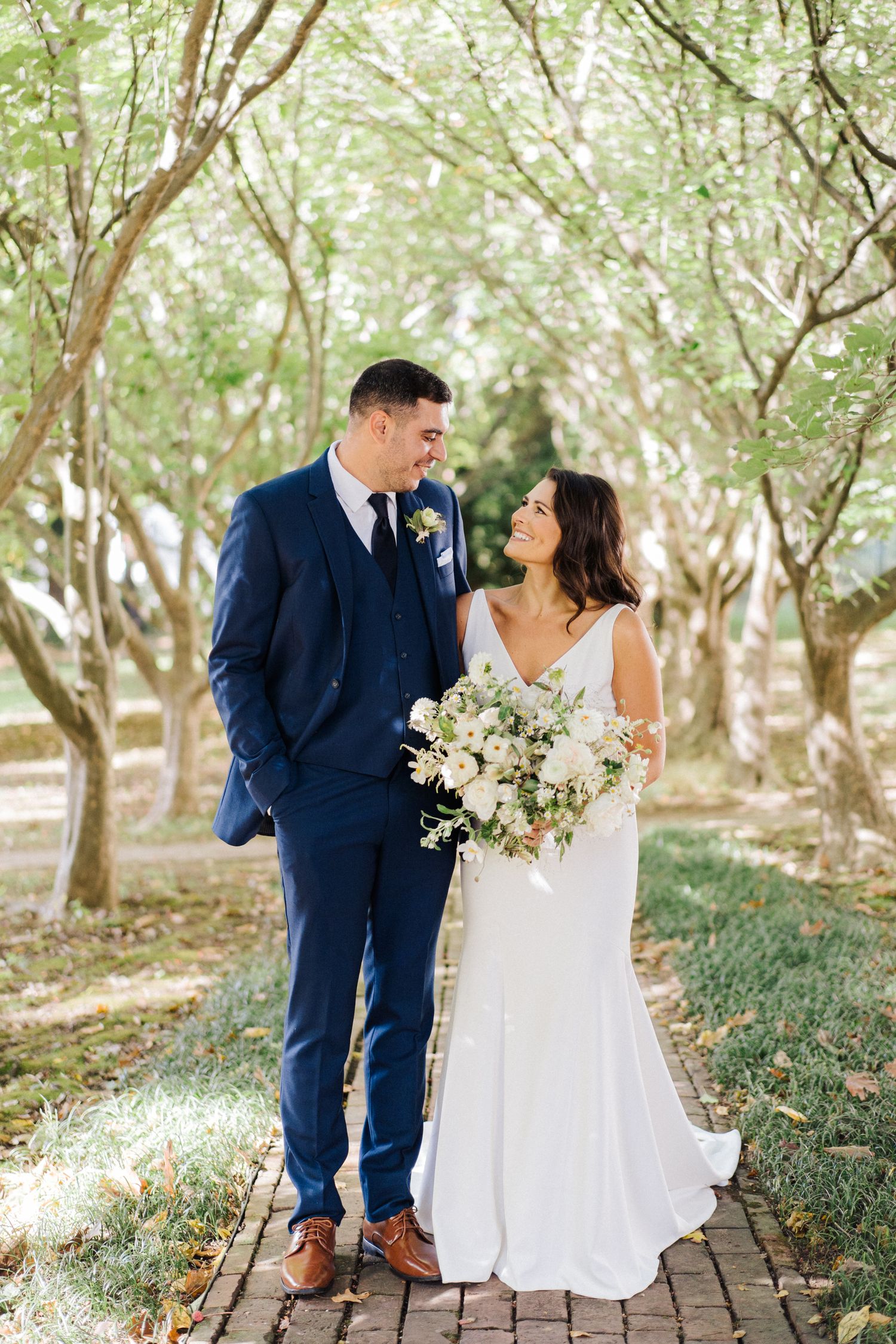 Elegant couple shares loving glance on brick pathway surrounded by trees.