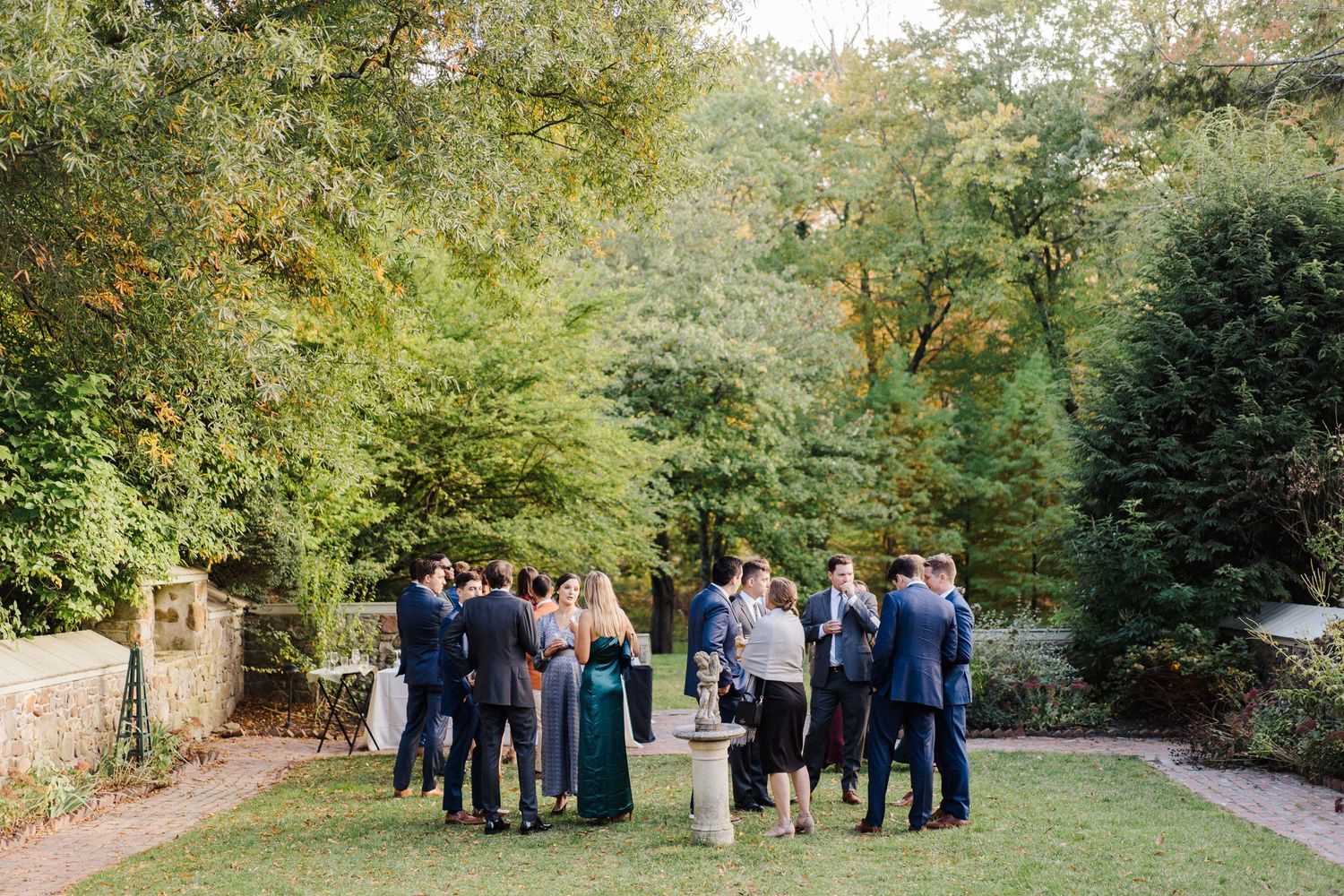 Wedding guests gather in a lush garden setting surrounded by green trees and foliage.