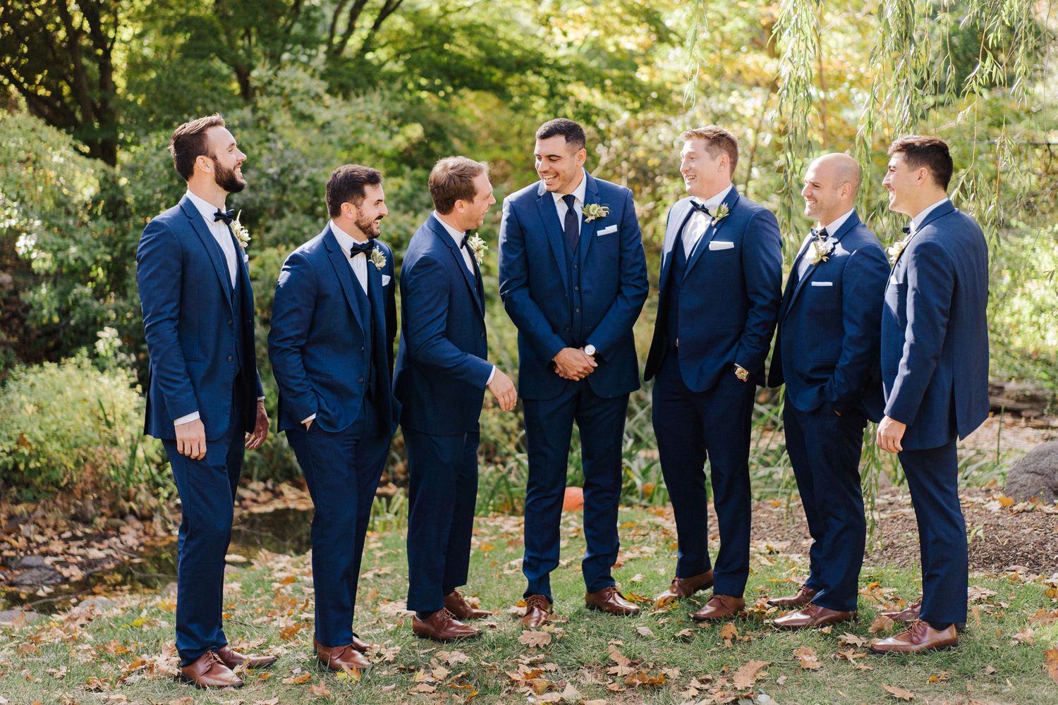 Wedding party in matching navy suits gather for outdoor photo beneath autumn trees.
