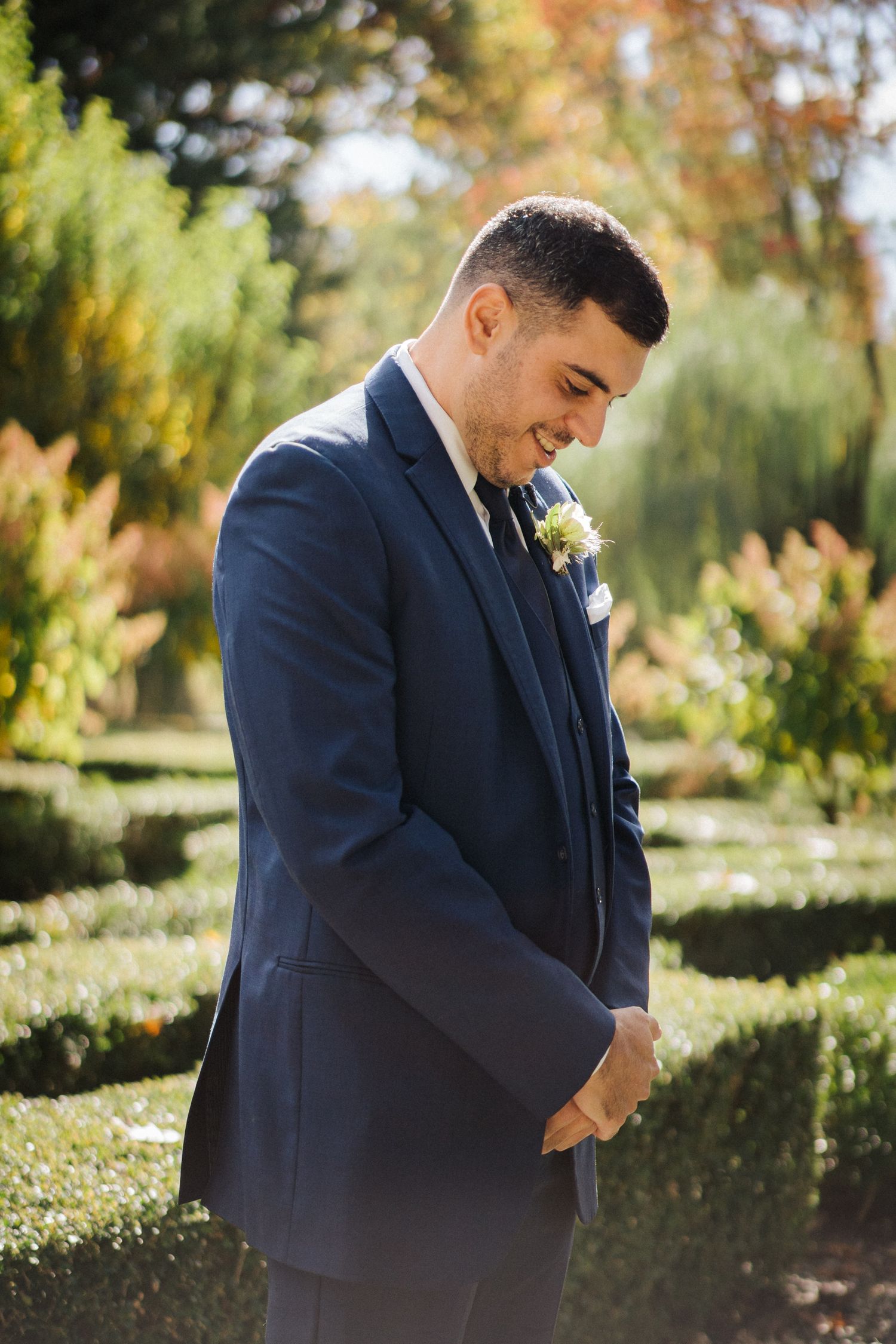 Groom in navy suit smiling in formal garden setting.