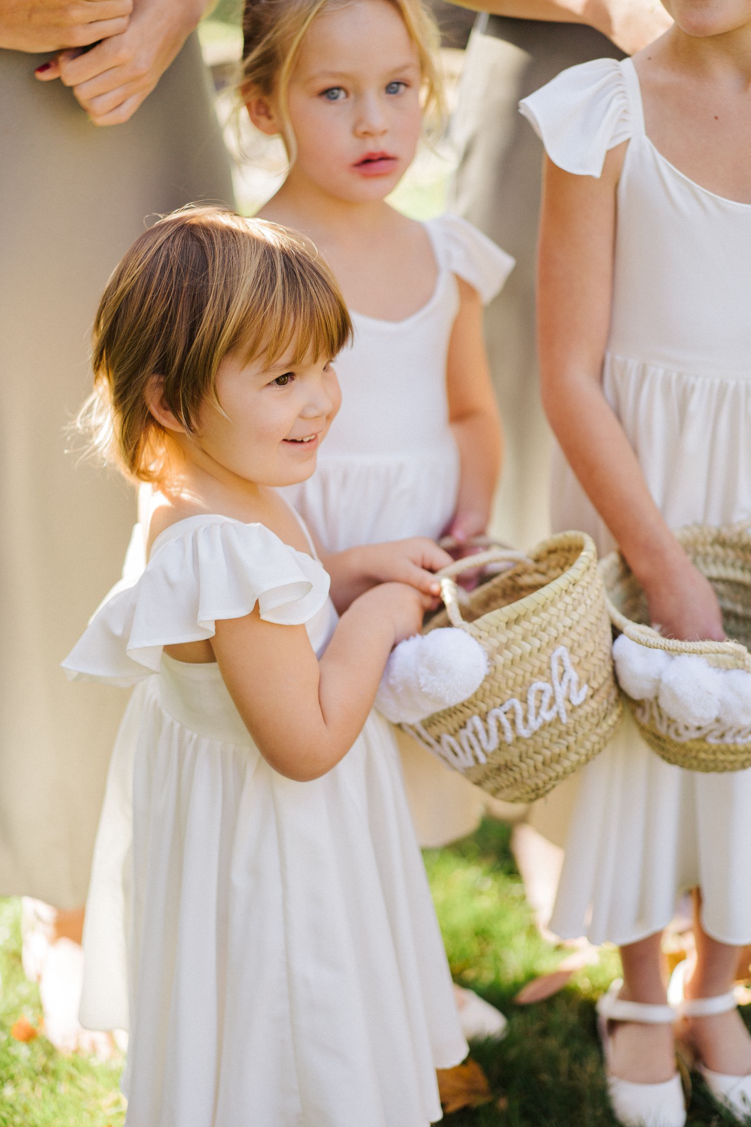 Young flower girls in white dresses hold decorated baskets during wedding ceremony.