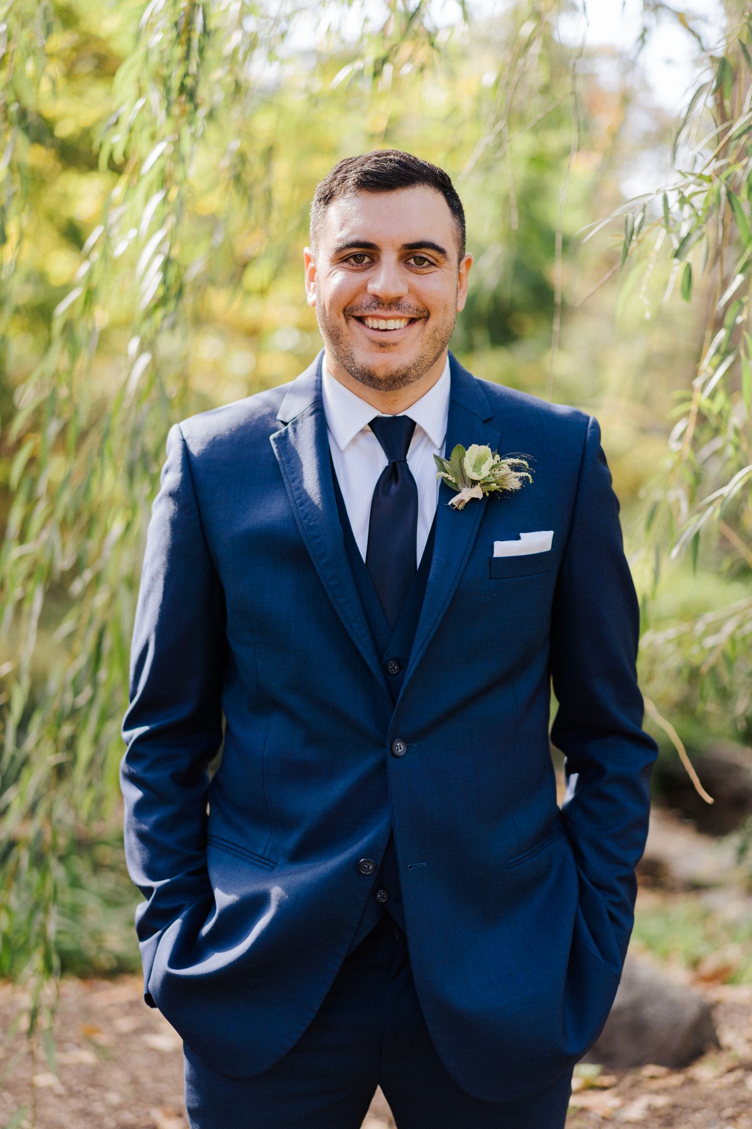 Groom in dark blue suit with boutonniere poses under willow trees.