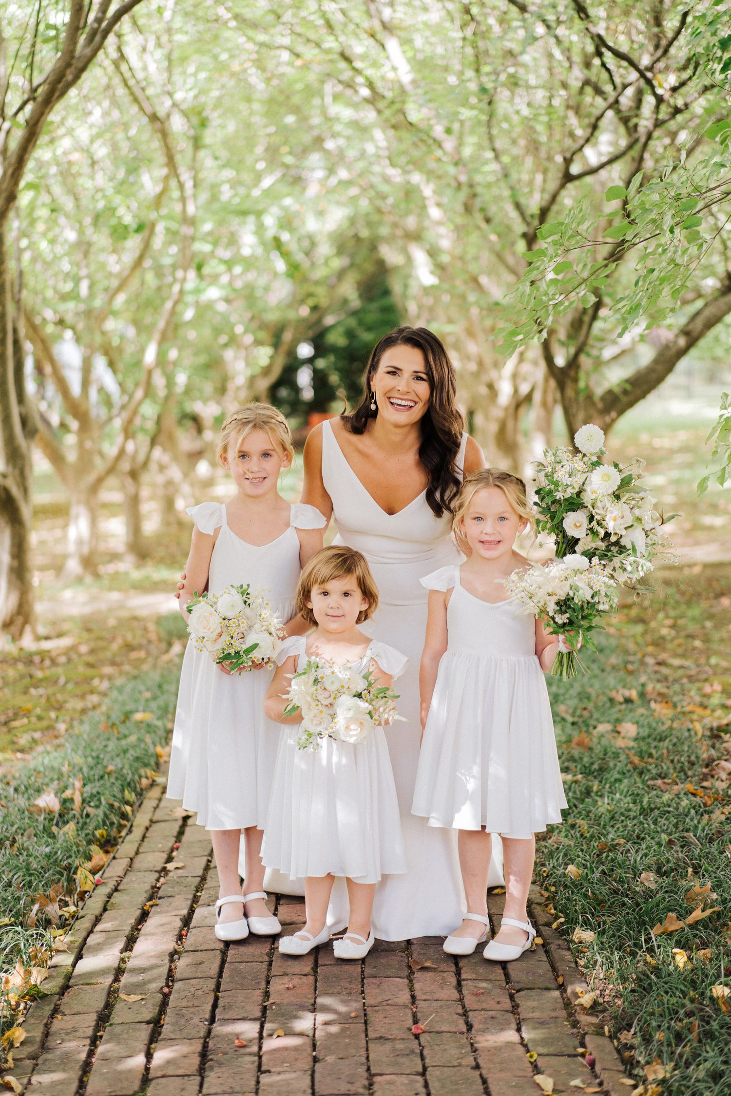 Group portrait on brick pathway with white dresses and floral bouquets in garden setting.
