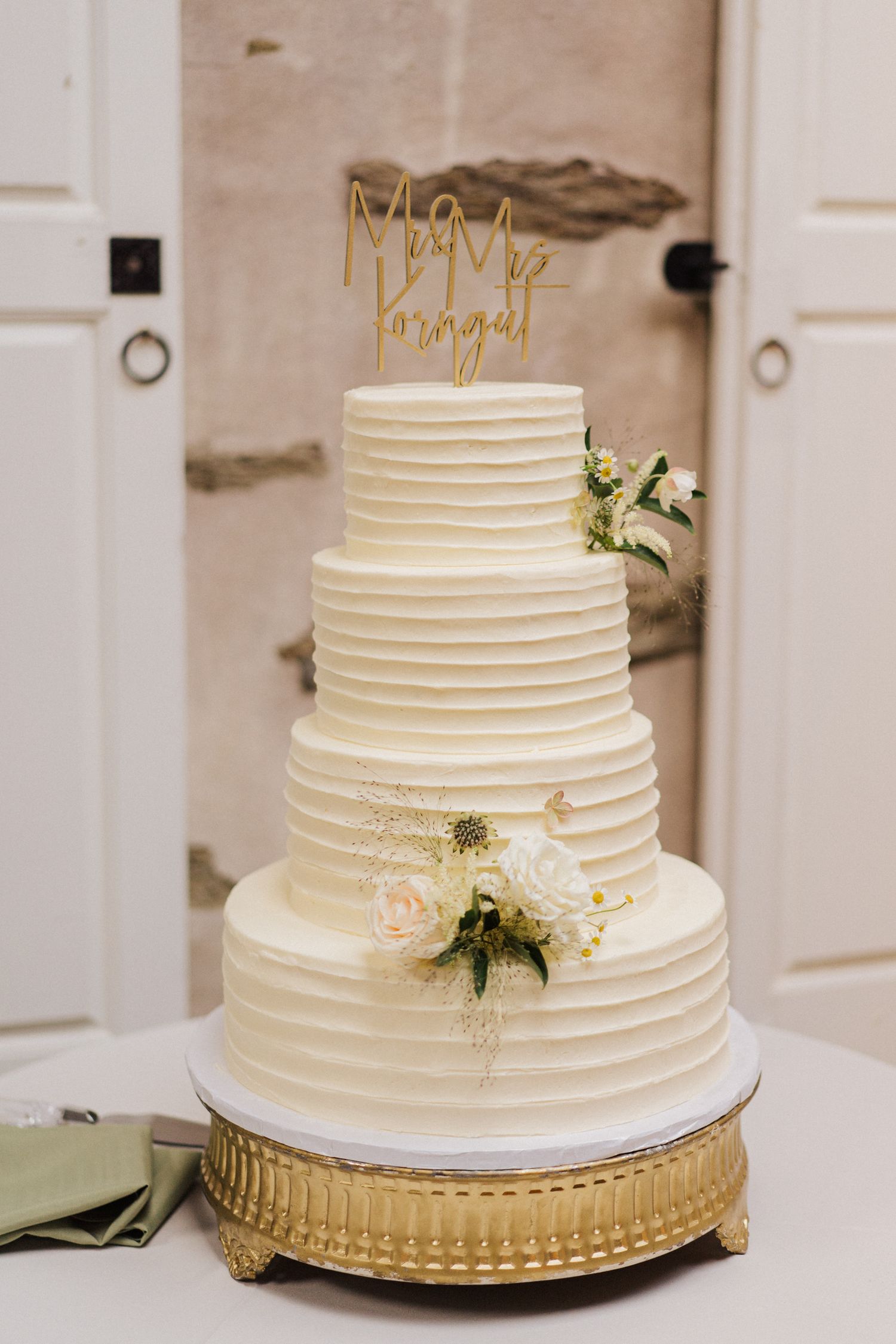 Elegant four-tier white wedding cake with horizontal lines and gold accents on ornate stand.