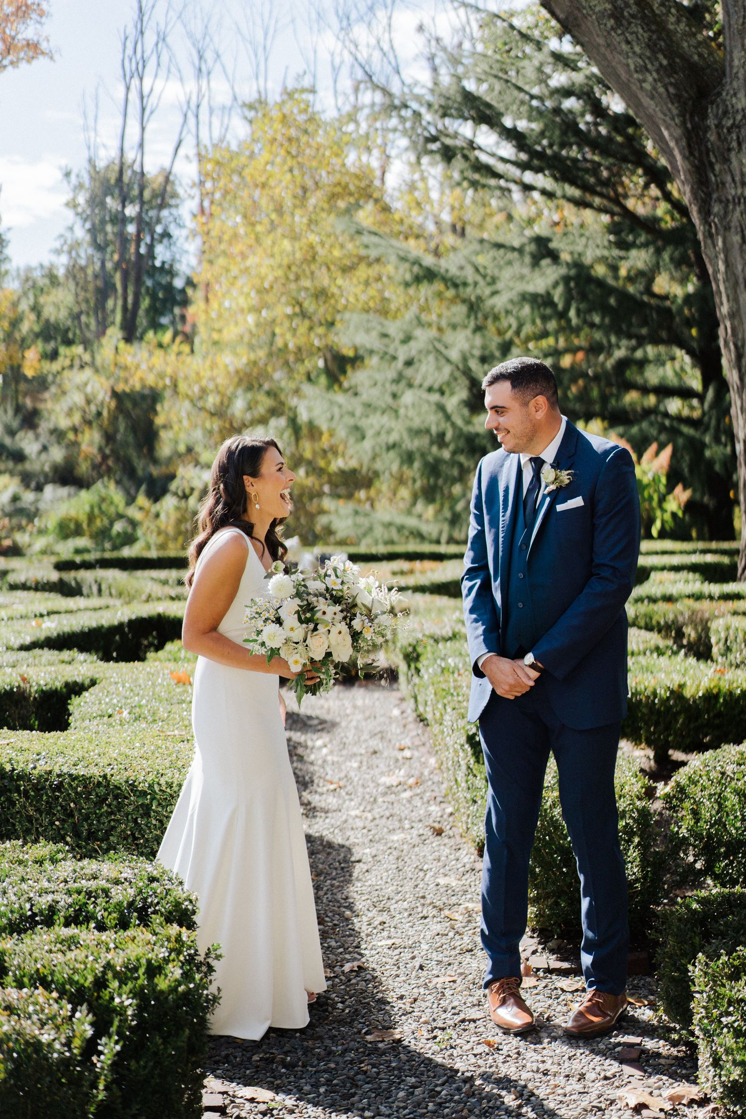 A romantic wedding scene on a gravel path with elegant landscaping and autumn trees in the background.