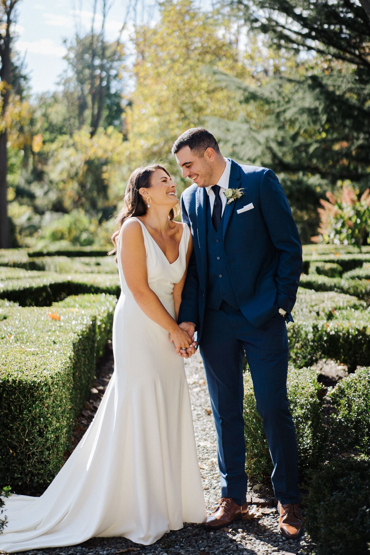 A candid wedding moment captured in a sunlit garden with geometric hedges and fall trees.