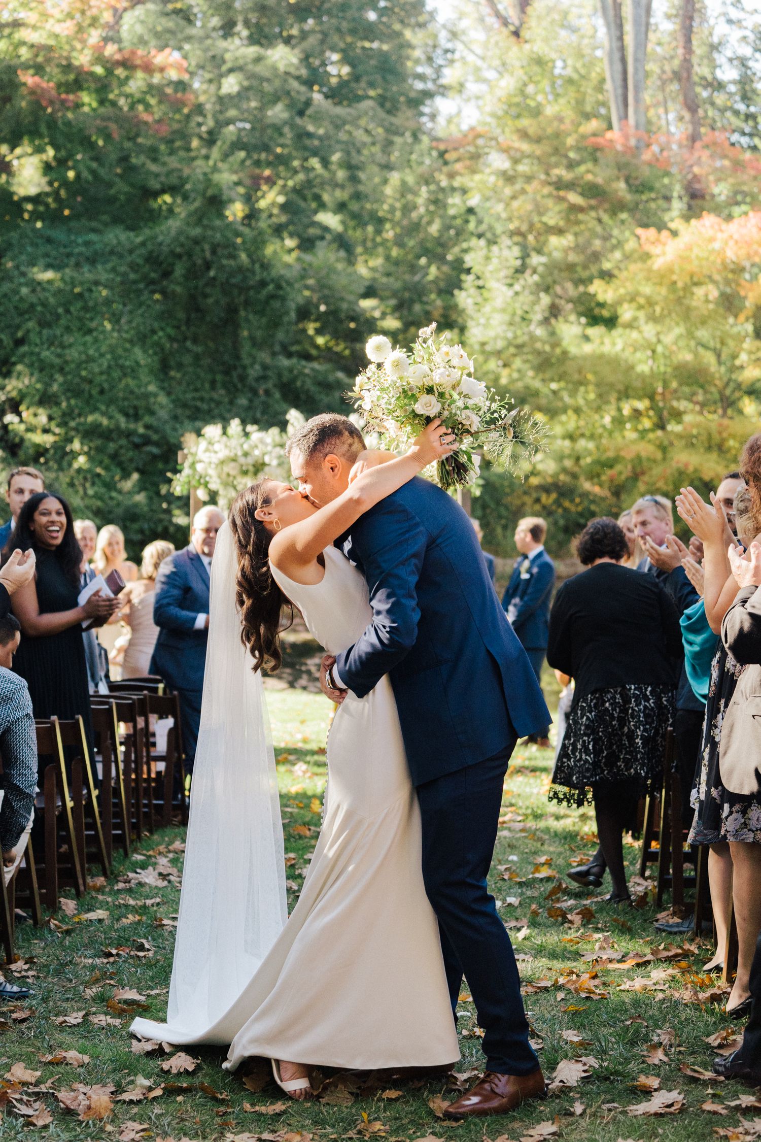 Newlyweds share their first dance on fallen leaves as guests applaud at outdoor reception.