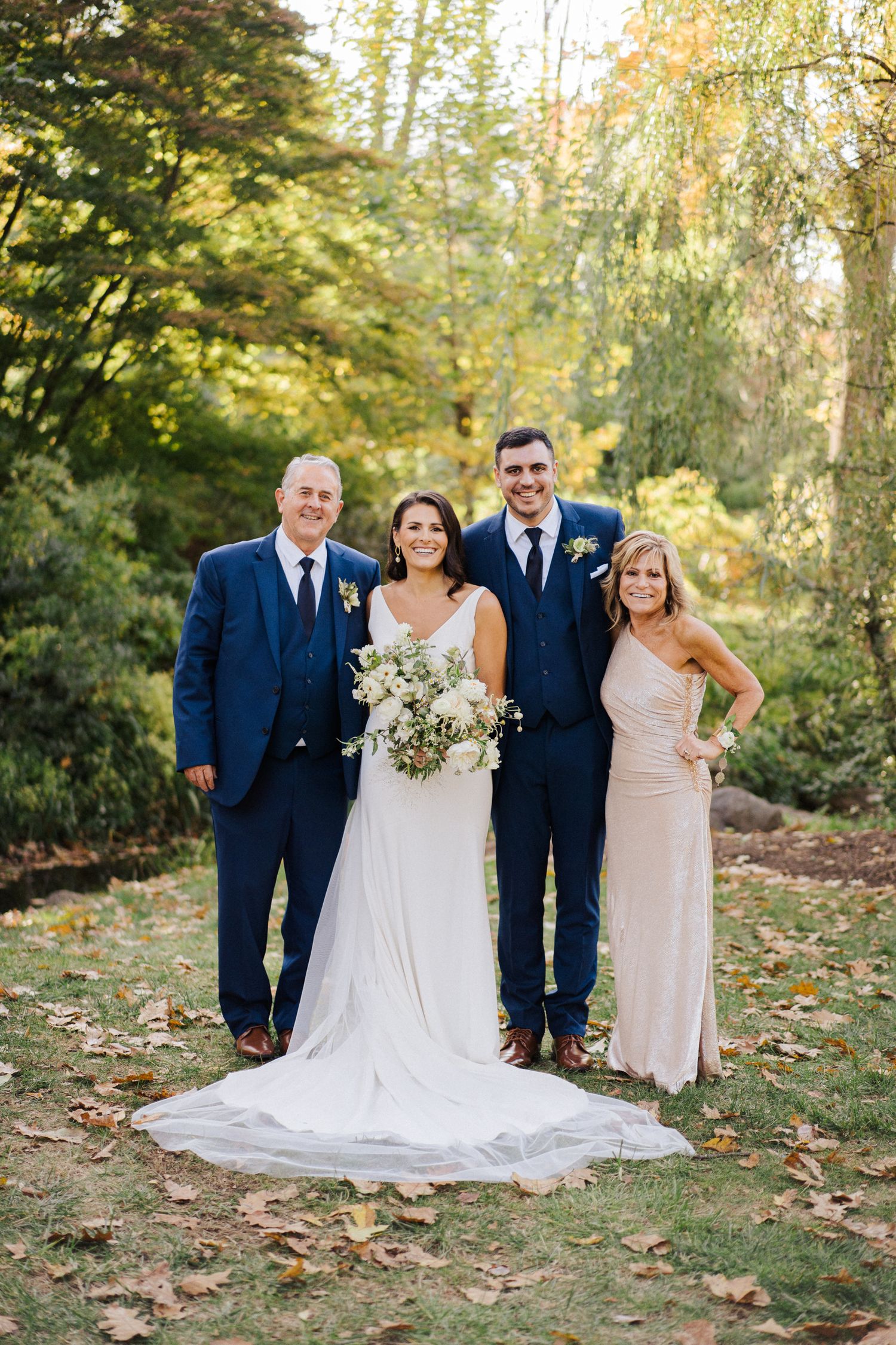 Wedding party in formal attire poses outdoors surrounded by autumn foliage.