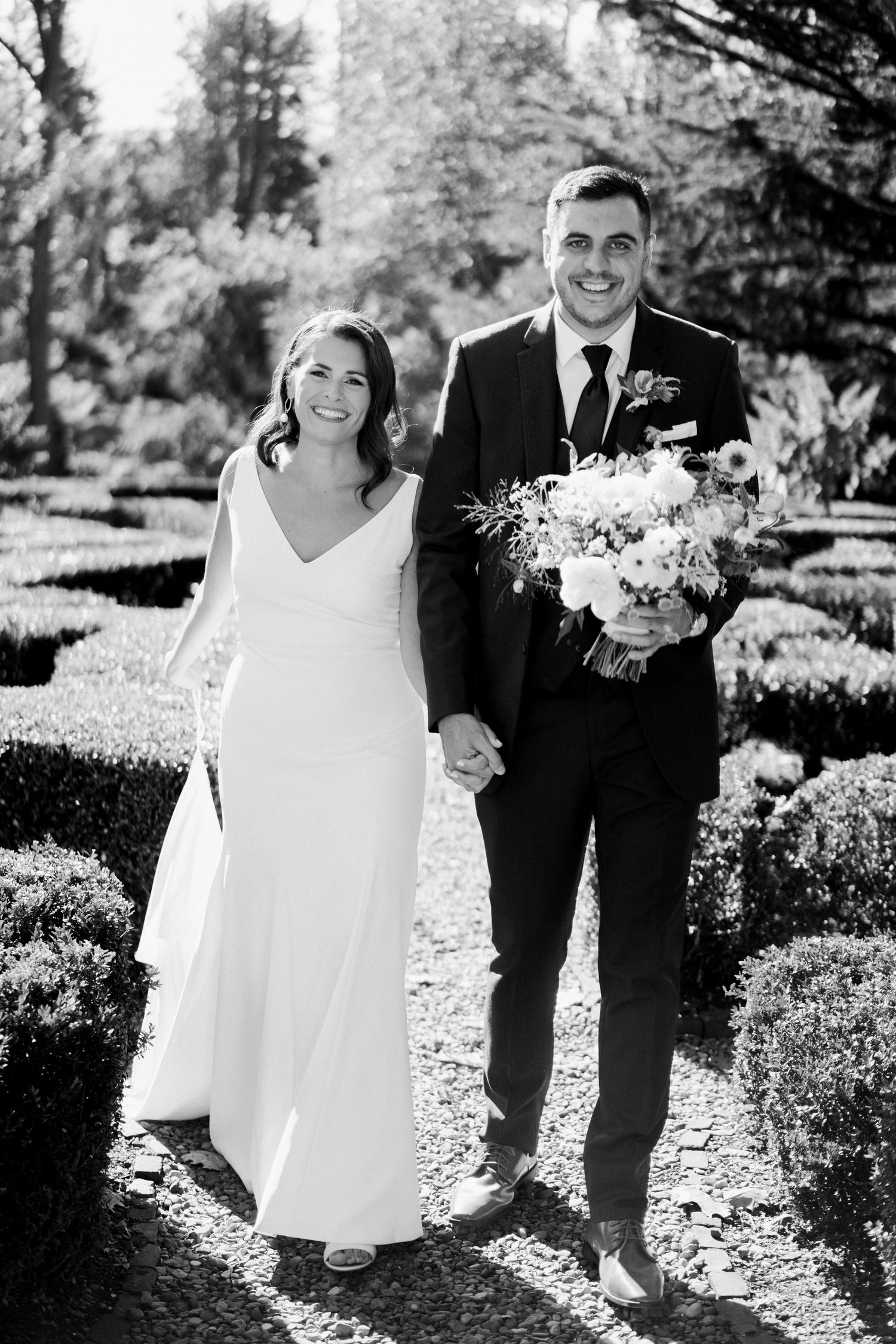 Black and white portrait of newlyweds holding hands on garden path with floral bouquet.