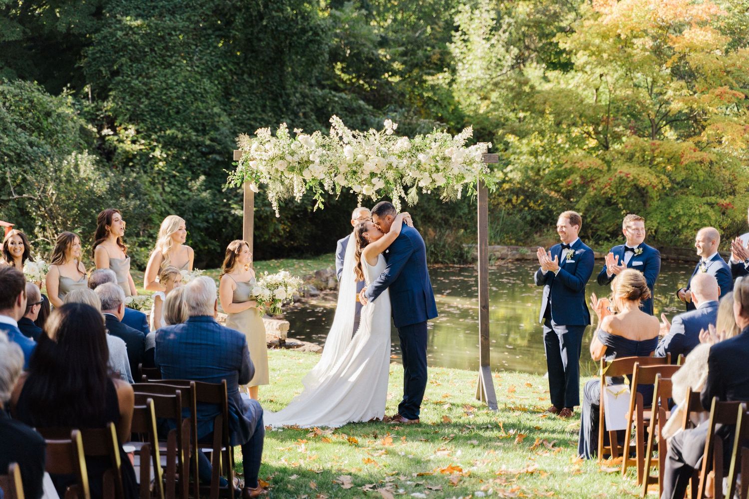 A wedding ceremony taking place in a garden setting with white floral decorations and wooden chairs.