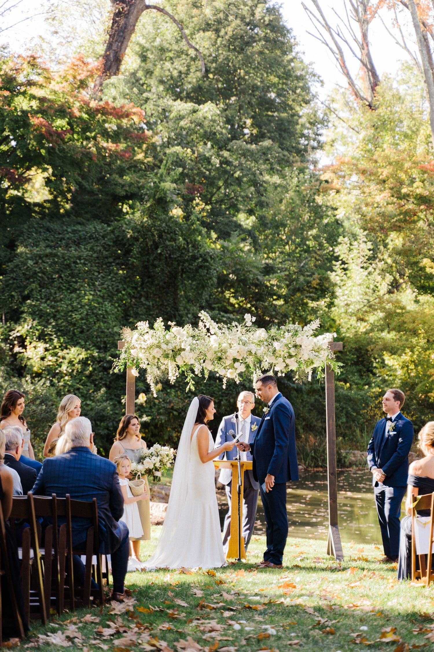 An outdoor wedding ceremony under a floral arch with guests seated on wooden chairs.