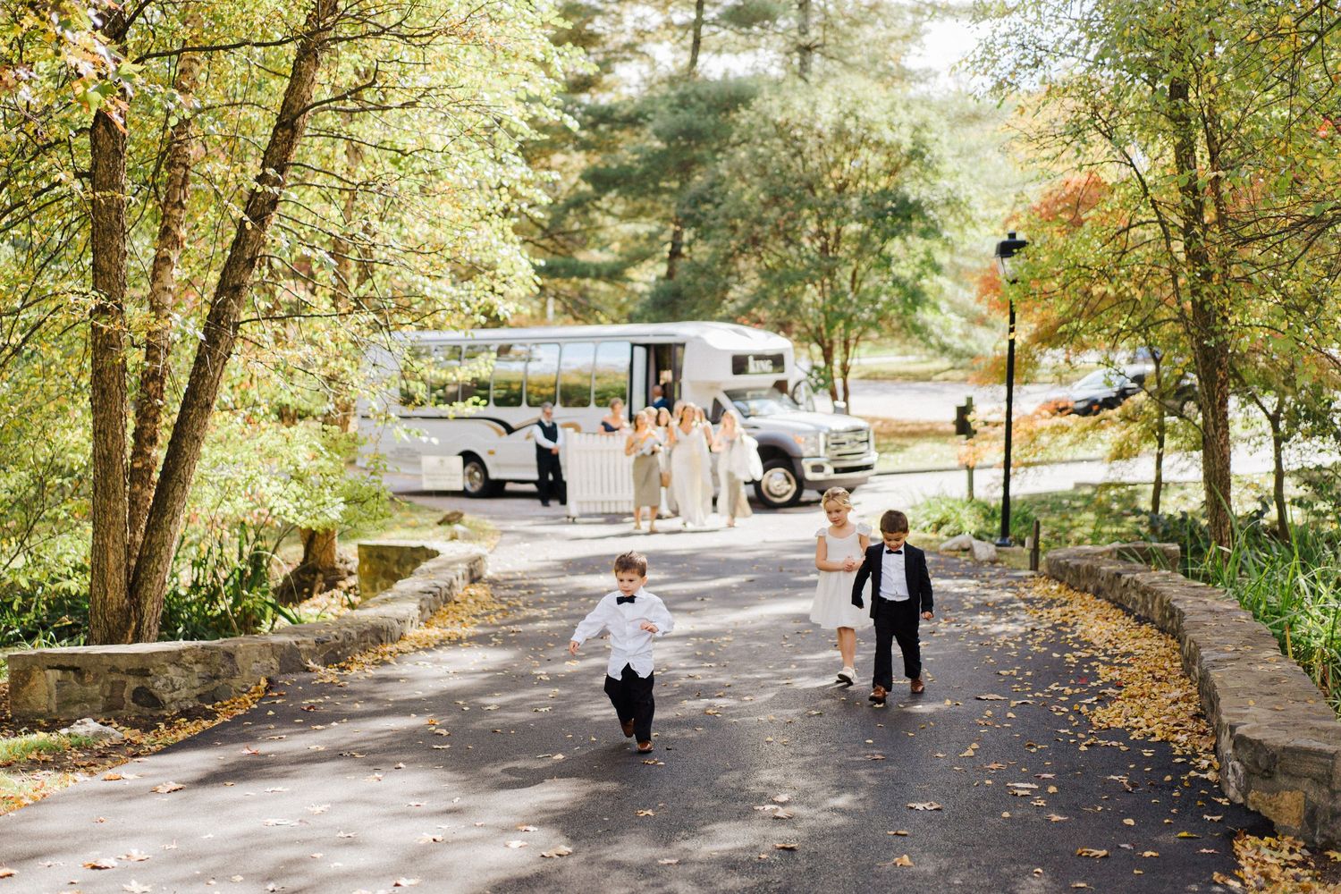 Wedding guests walk down a tree-lined path towards a white tent venue on an autumn day.