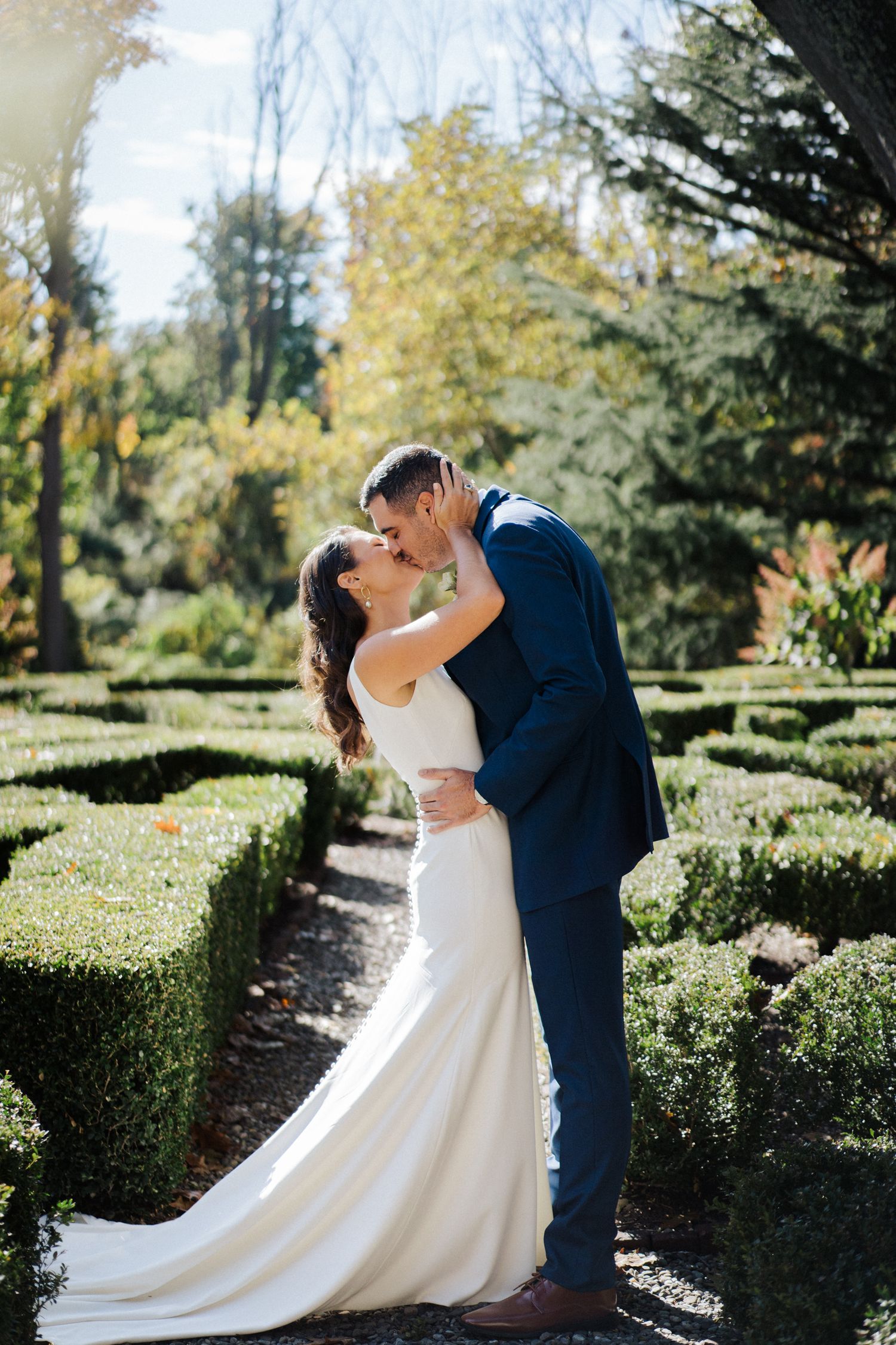 Romantic wedding couple sharing an intimate moment in a manicured garden with hedges on a sunny day.