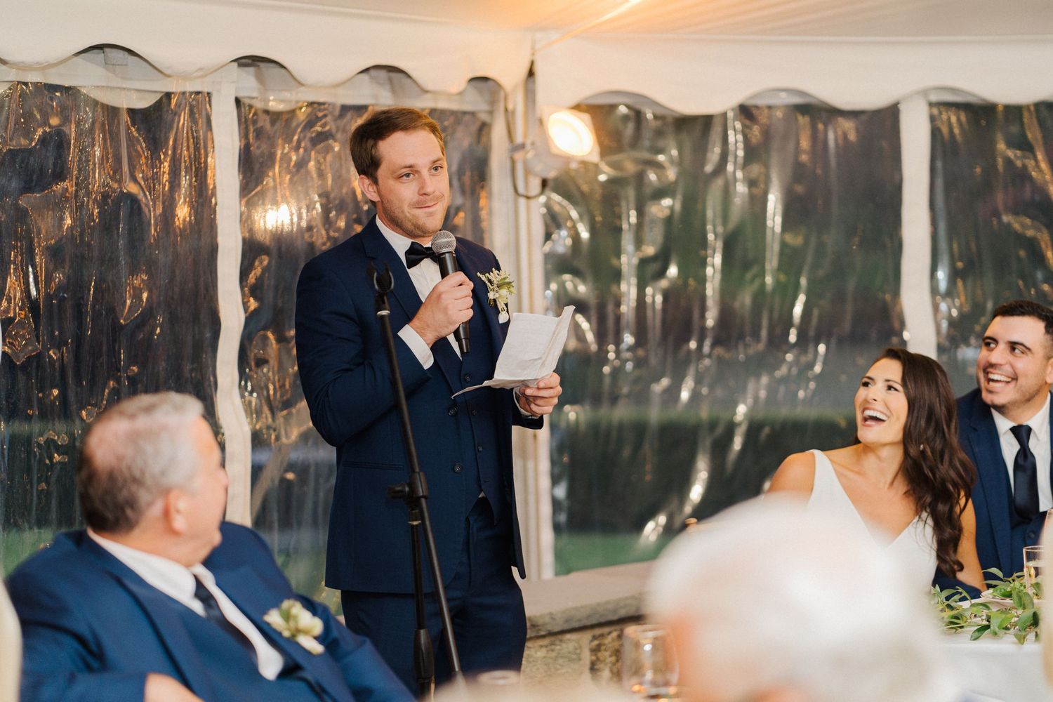 A speaker delivers a humorous wedding toast as guests laugh during the reception.