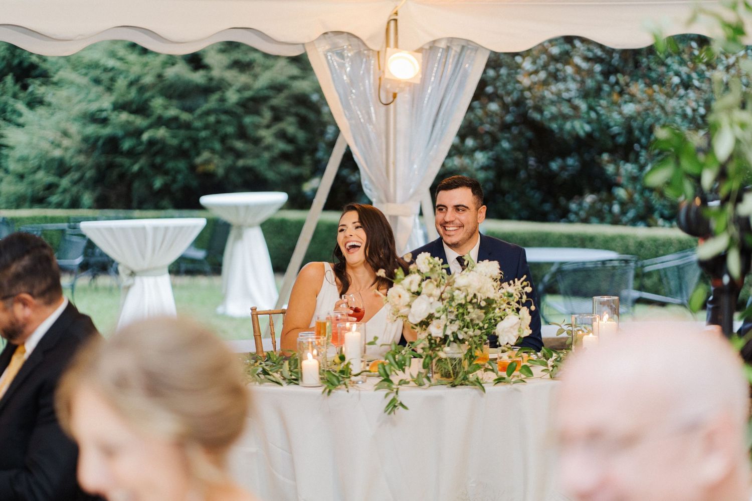A couple laughs together at their reception head table decorated with white linens and greenery.