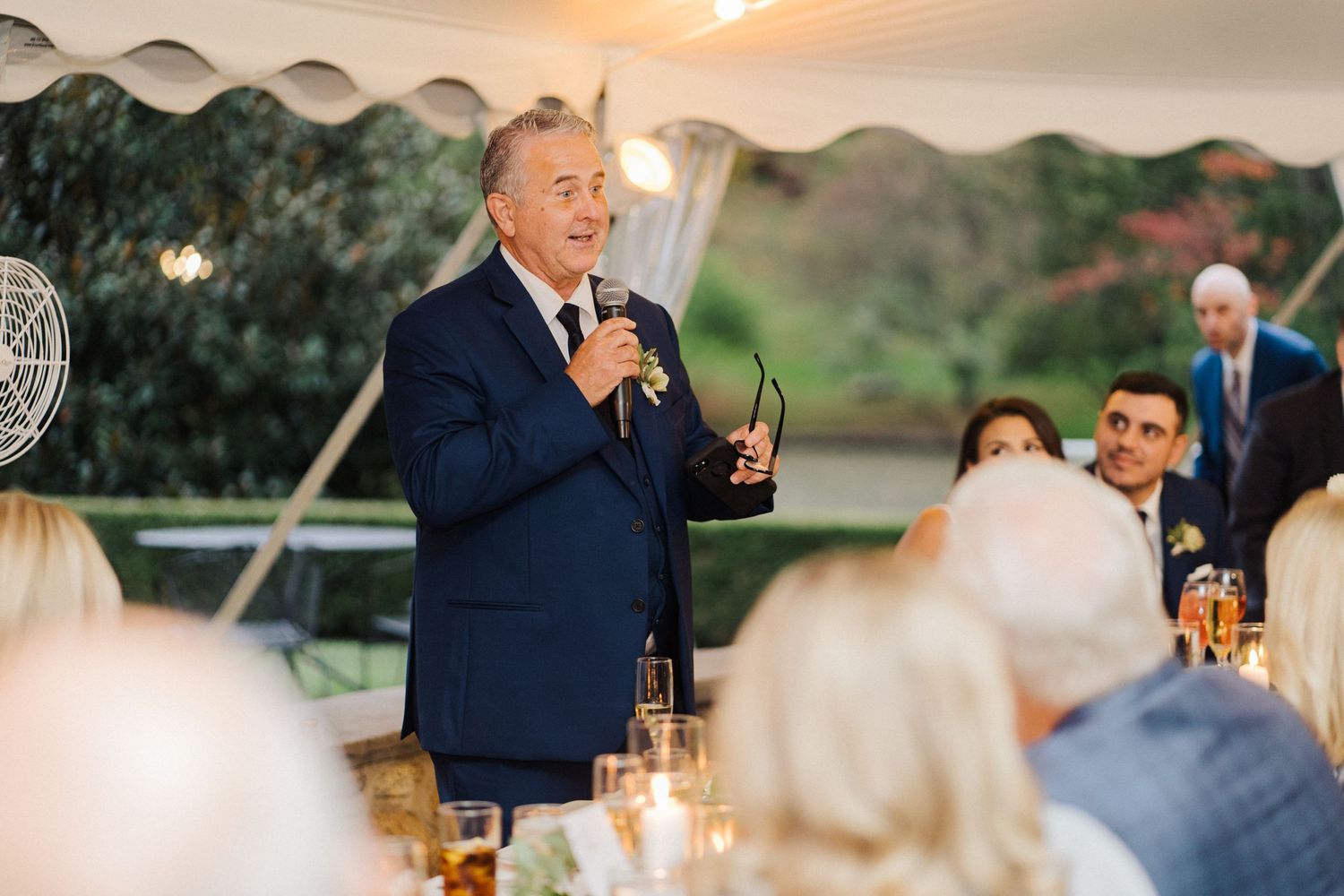 A guest gives a toast during an outdoor wedding reception under string lights at sunset.