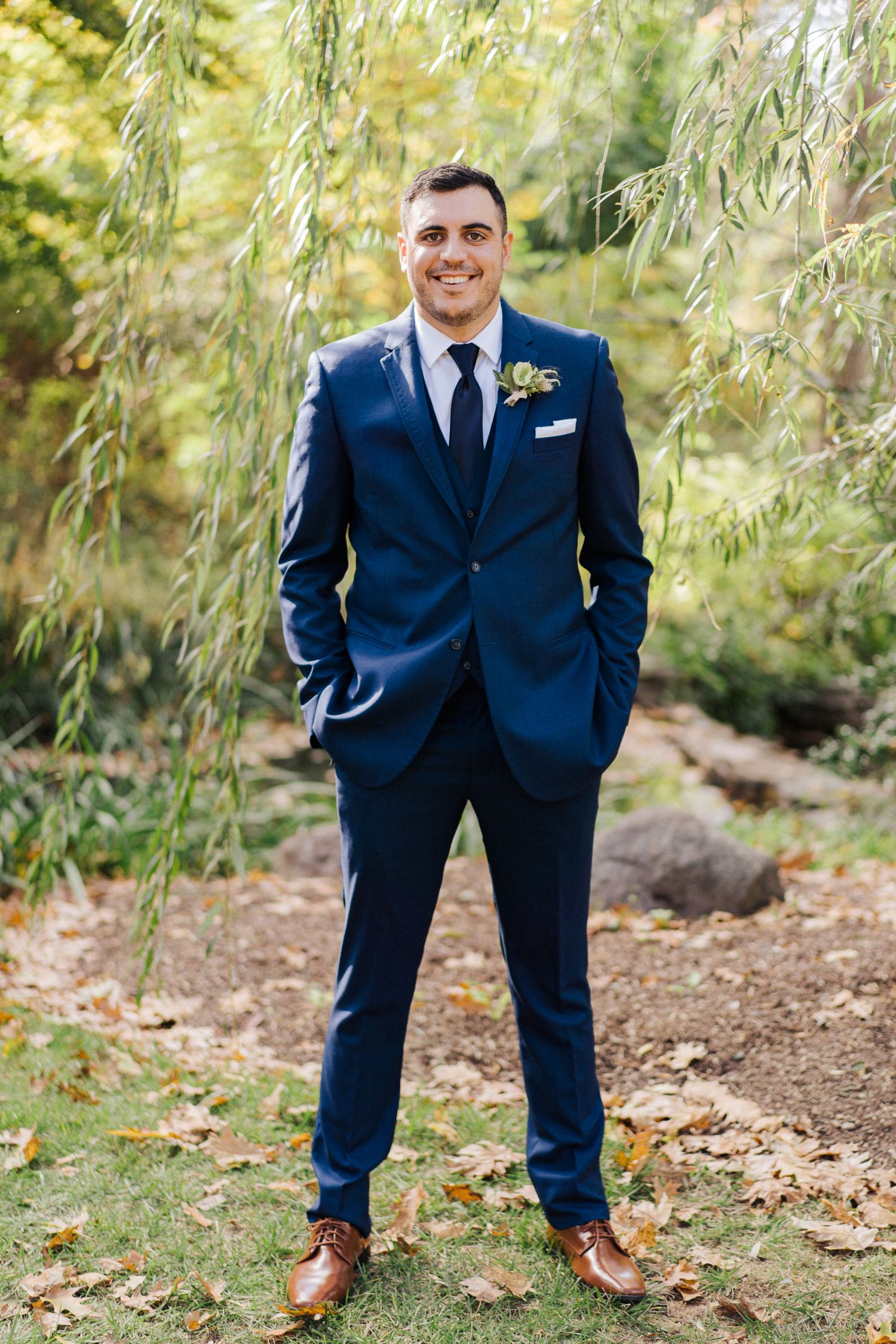 Groom in navy blue suit and brown shoes stands confidently under willow trees.