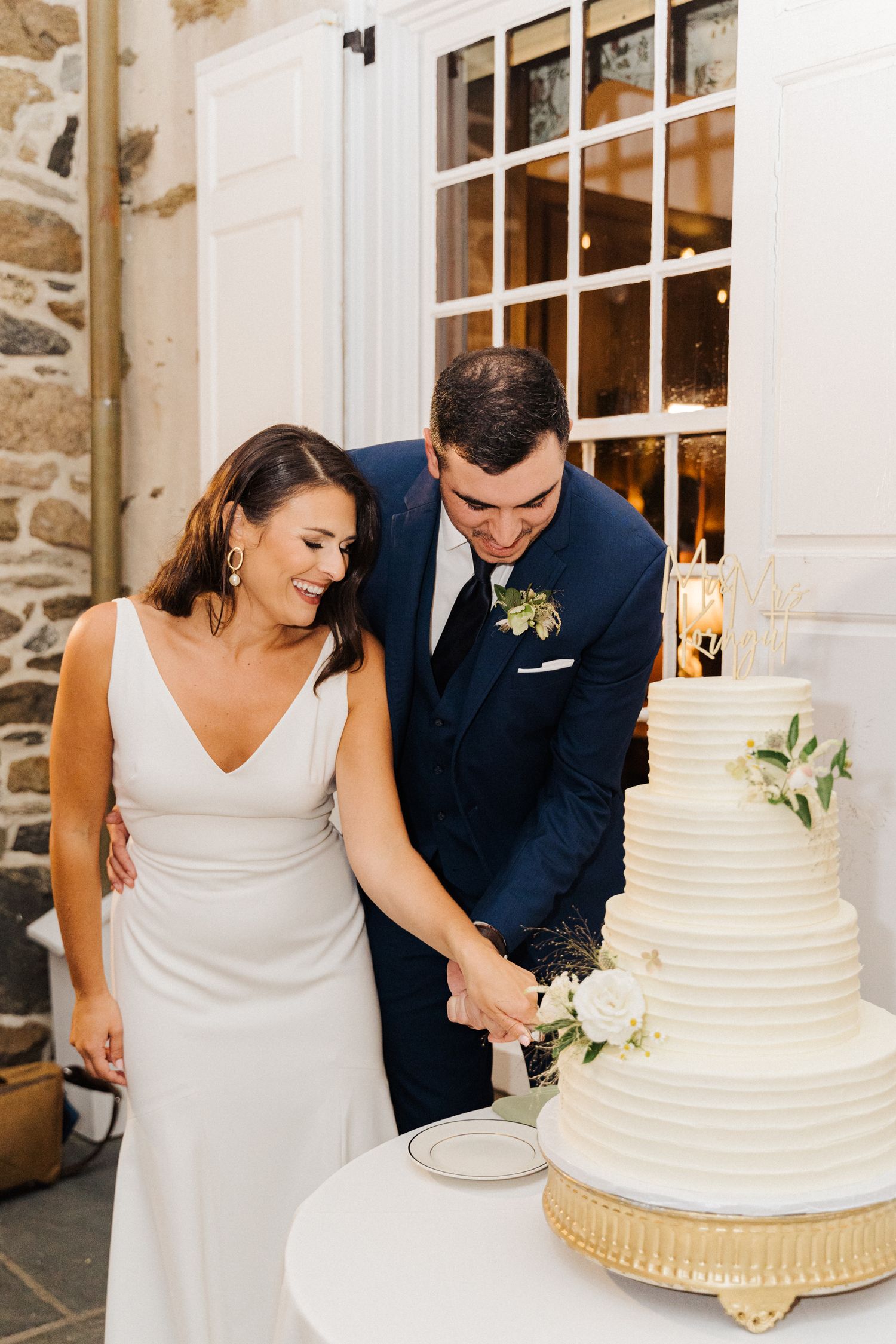 A couple cuts their elegant white tiered wedding cake together by a stone wall.