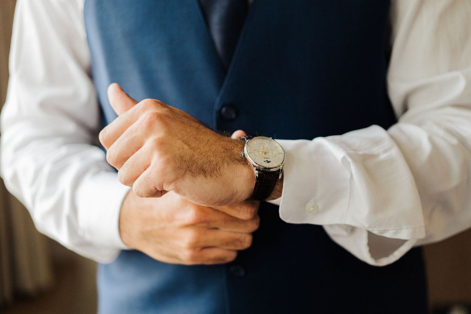 Close-up of hands adjusting cufflinks on white dress shirt with blue vest.