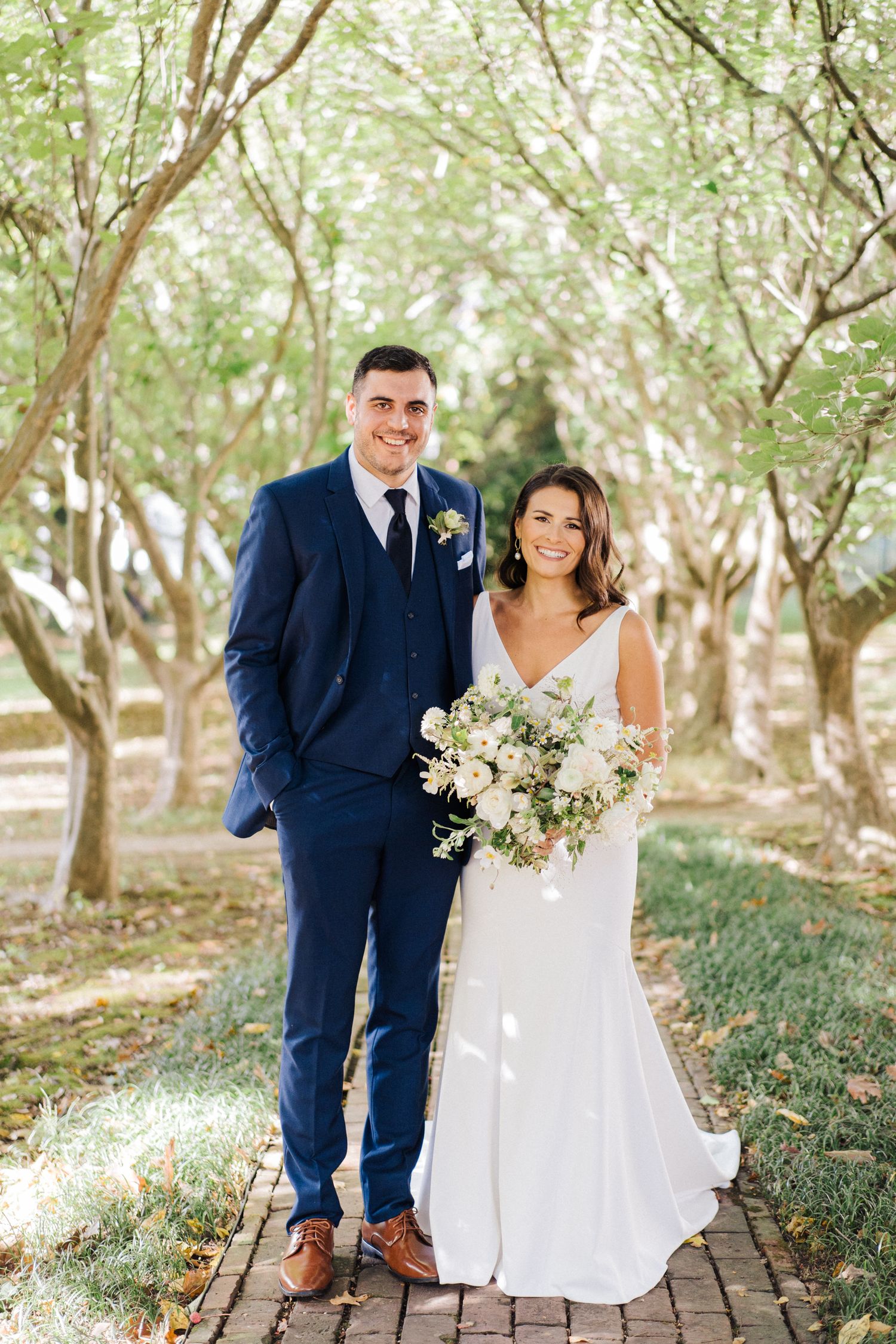 Wedding couple poses on brick pathway under canopy of trees in romantic garden setting.